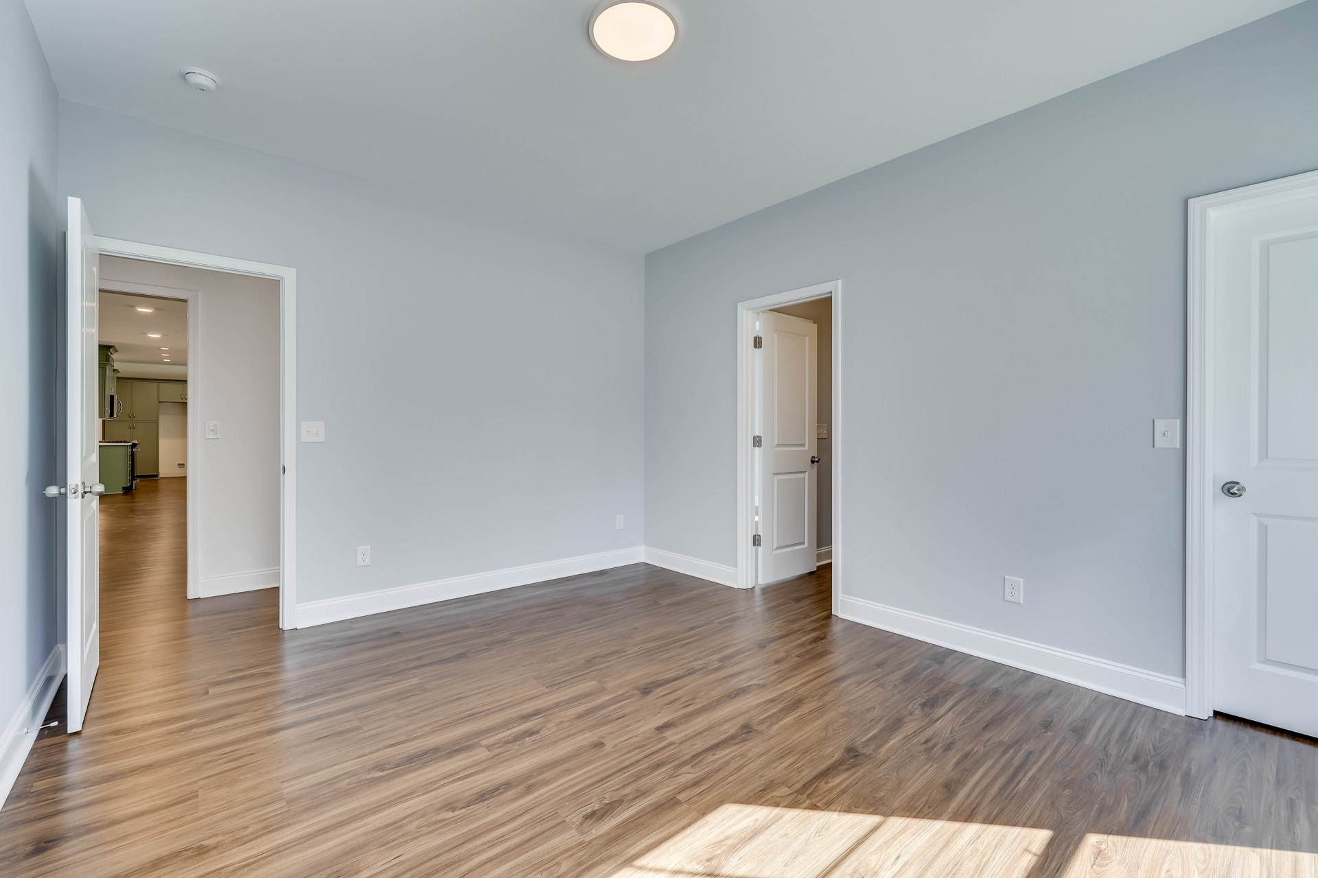 Room with light wood flooring, white walls, and a white door featuring a silver knob; round white ceiling light fixture visible.