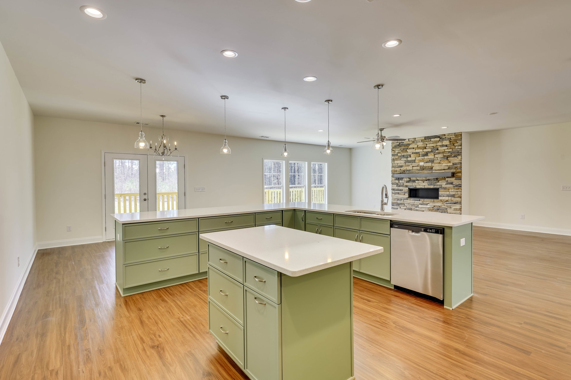 Spacious kitchen featuring a large island with white countertop, wood flooring, built-in drawers and cabinets, stainless steel dishwasher, and a window overlooking a forest.