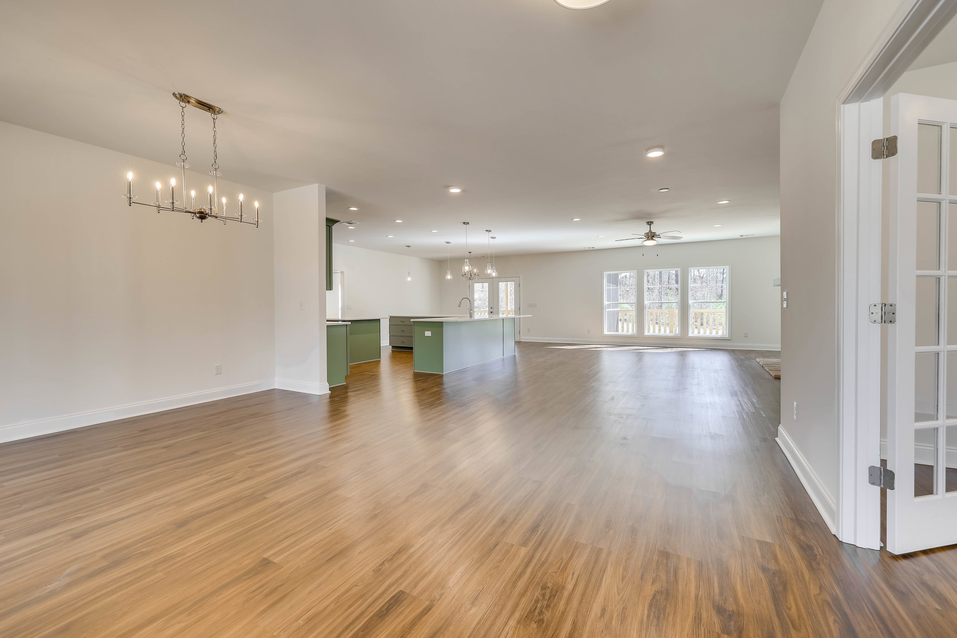Spacious open-plan room with hardwood flooring, ceiling fan, chandelier, white kitchen countertops, large window with railing, and view of trees outside