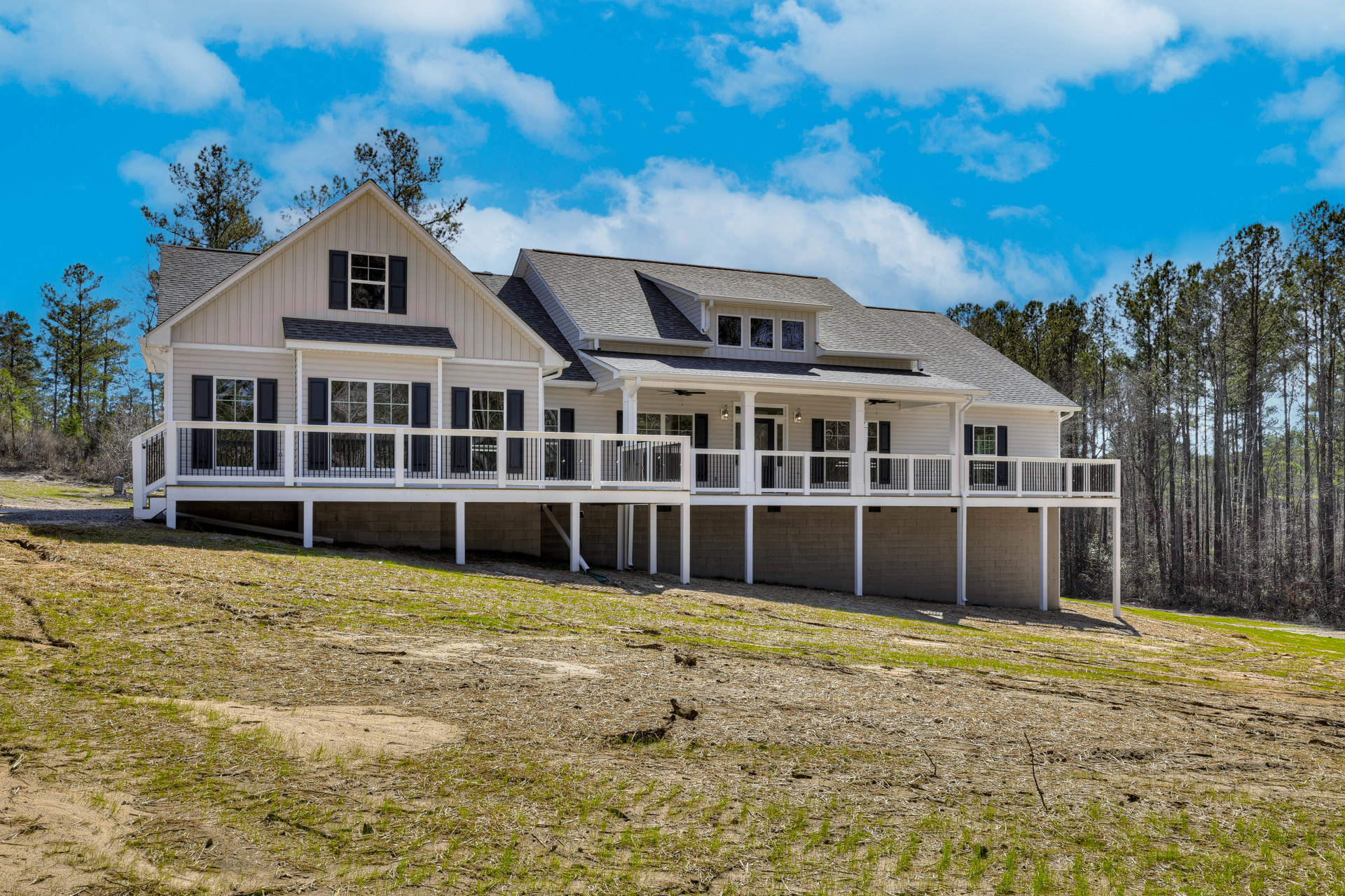 White house with expansive deck featuring black railings, set on a grassy lawn with mature trees and blue sky in the background