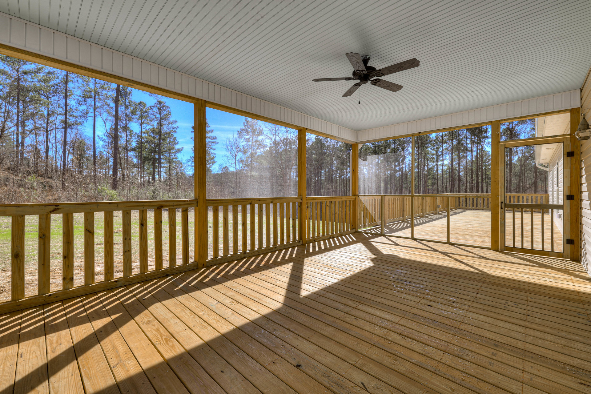 Wood deck with water droplets, ceiling fan with wood blades, partial porch roof, fence, and leafy trees in the background