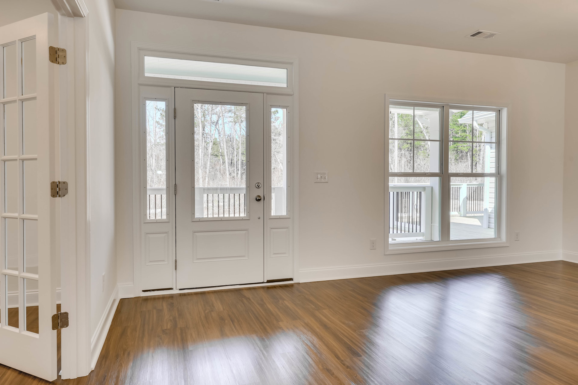 White walls and hardwood floor in a bright room, featuring a white double door with glass panes, a window overlooking a deck and trees, and a close-up of a door hinge.