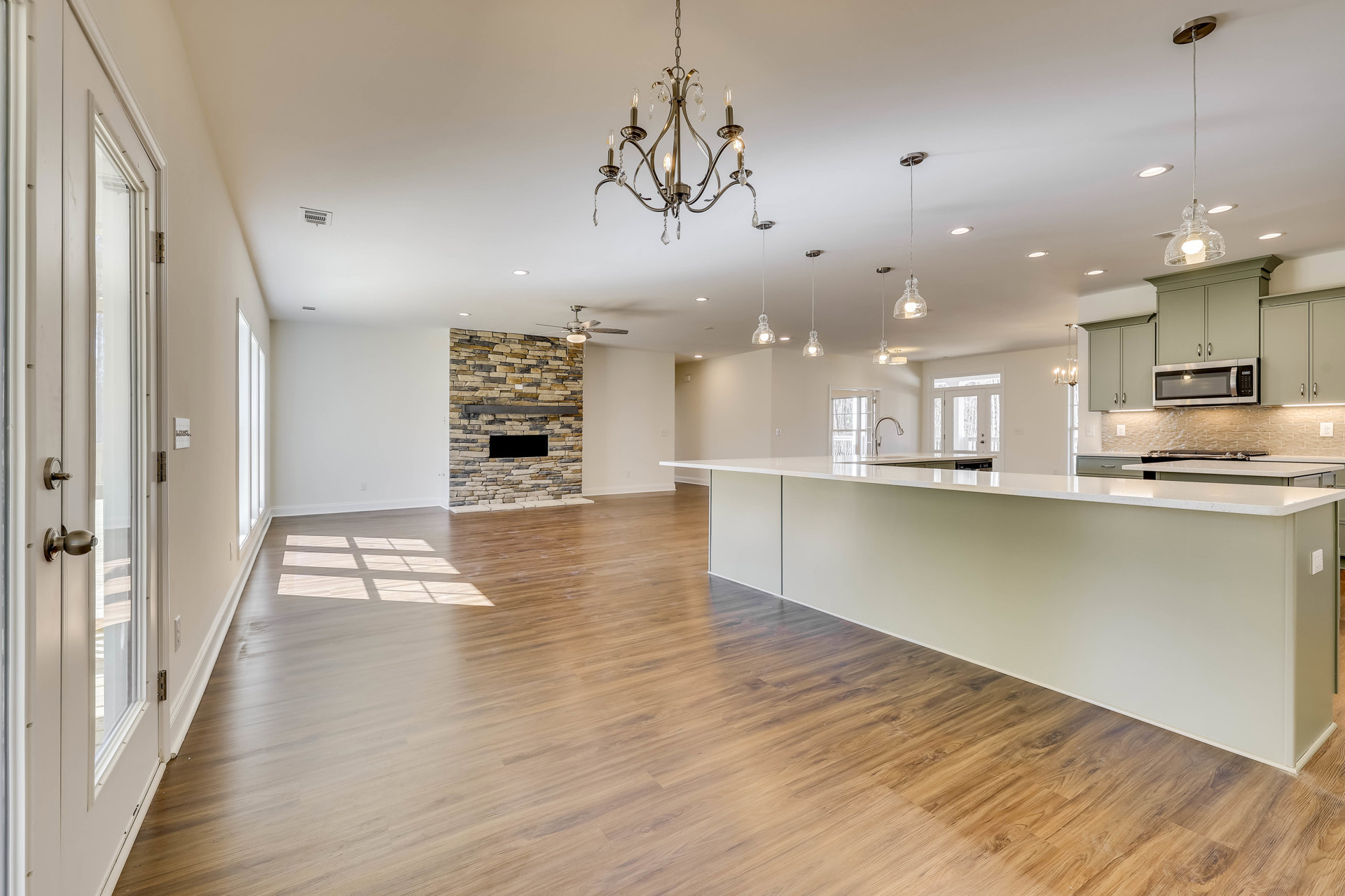 Spacious open room with wood flooring, stone fireplace with built-in shelf, modern kitchen featuring wood cabinetry, laminate countertops, stainless steel sink, and crystal