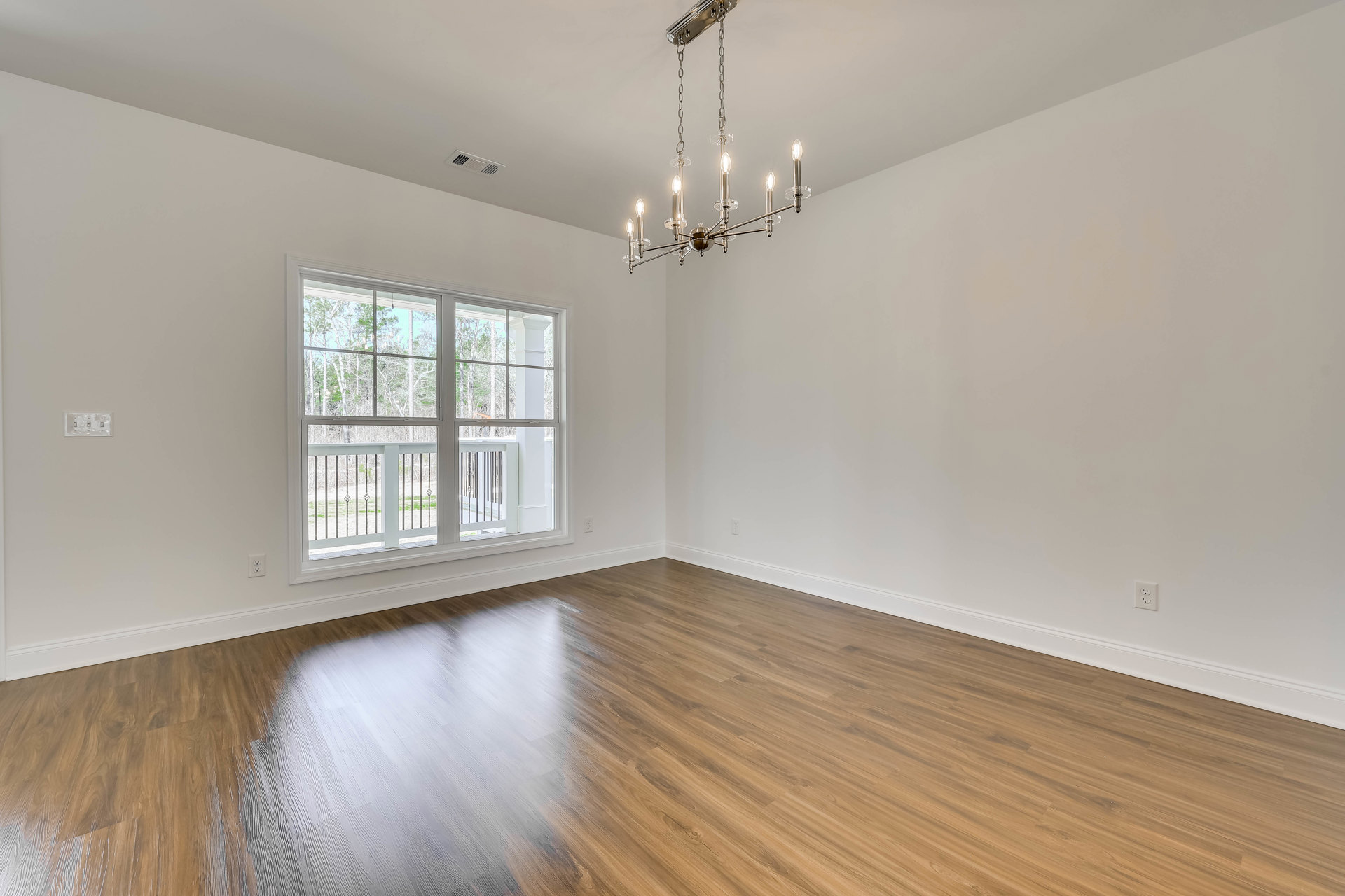 Wood flooring and white walls in a spacious room, ceiling-mounted chandelier with clear lights, large window with metal railing overlooking trees.