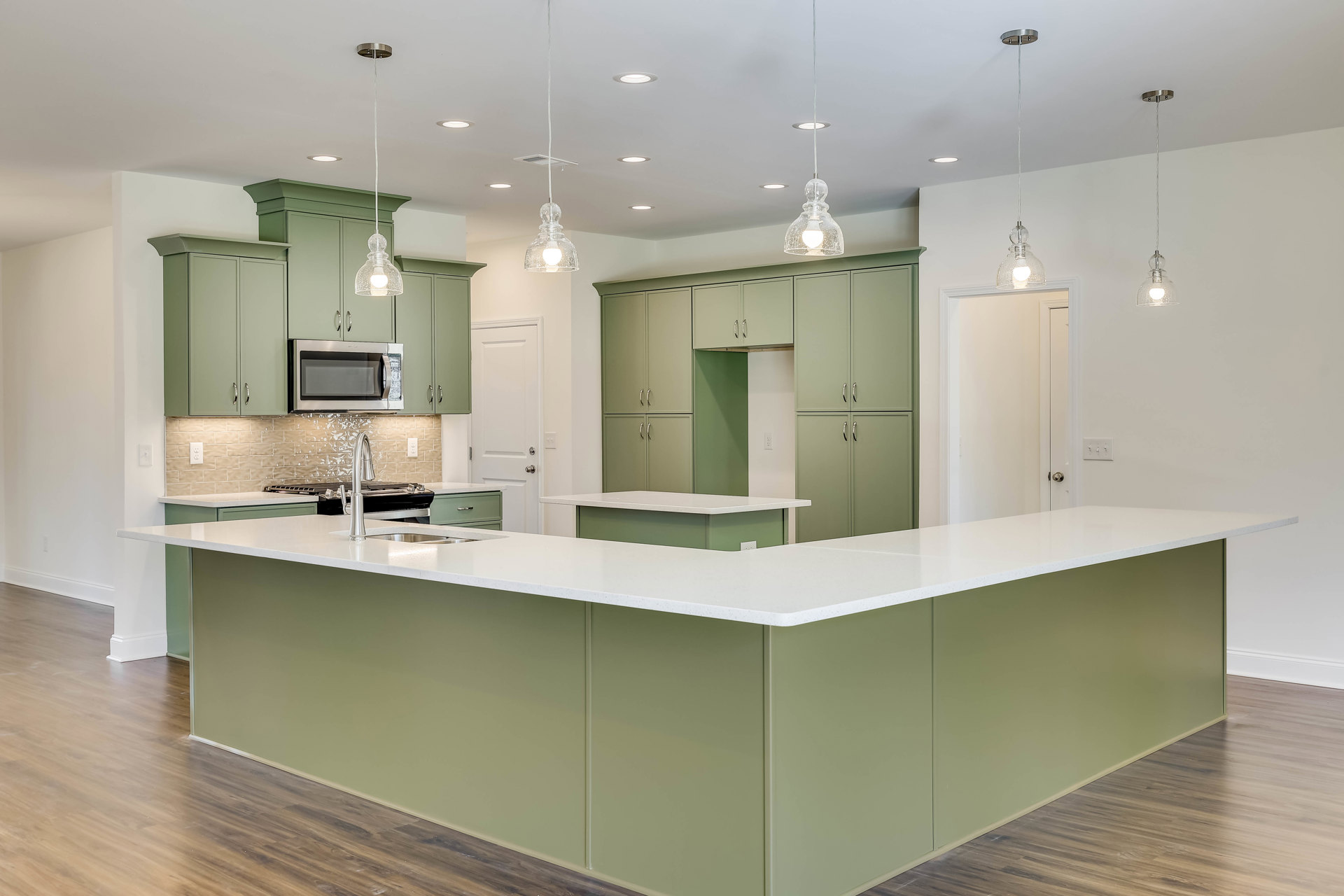 Green kitchen cabinets with white countertops, built-in microwave with glass door, ceiling vent, glass pendant light fixture, and slim hanging light above workspace.