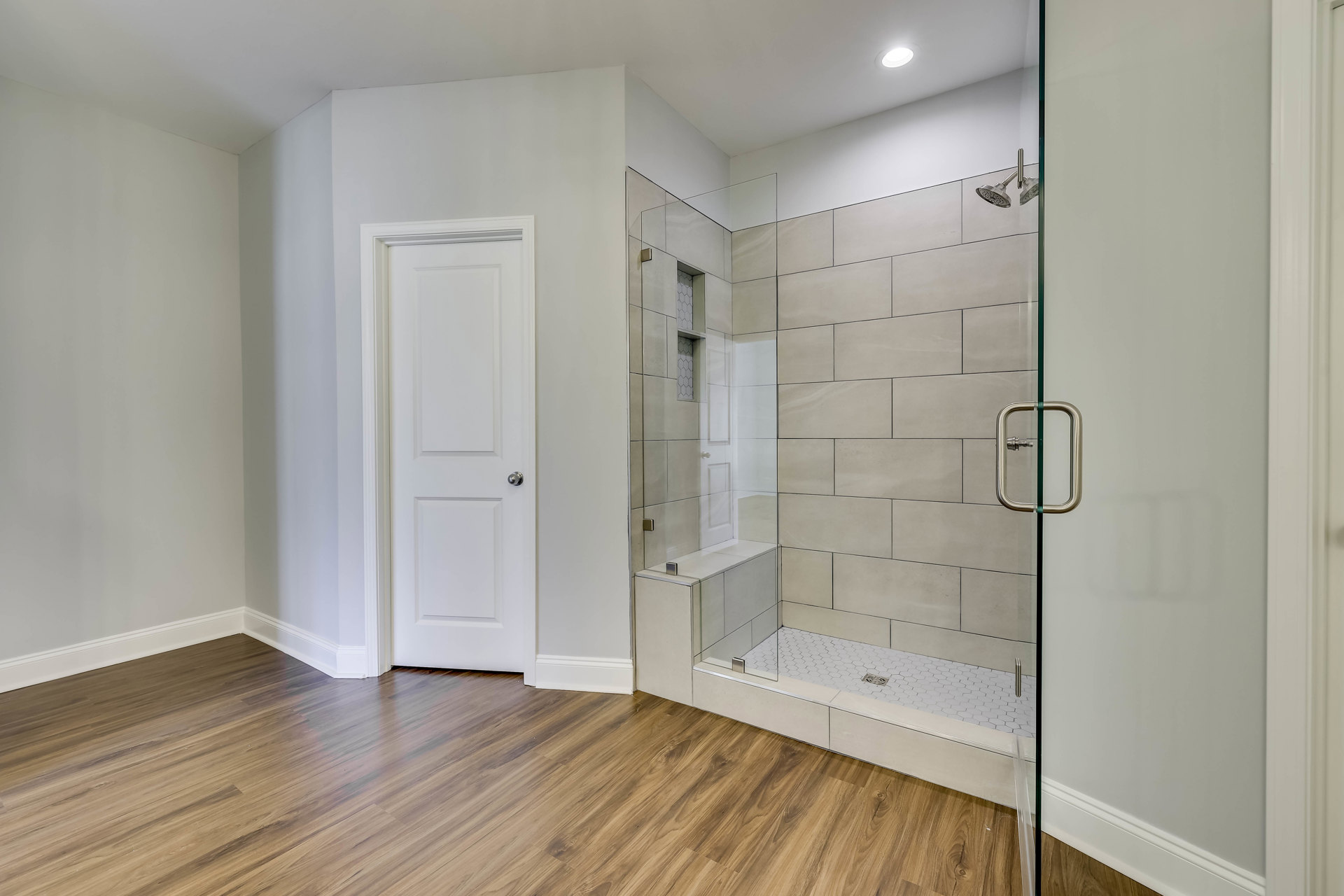 Bathroom with glass shower enclosure, white tile shower floor, wood plank flooring, white door with silver handle, white baseboard trim