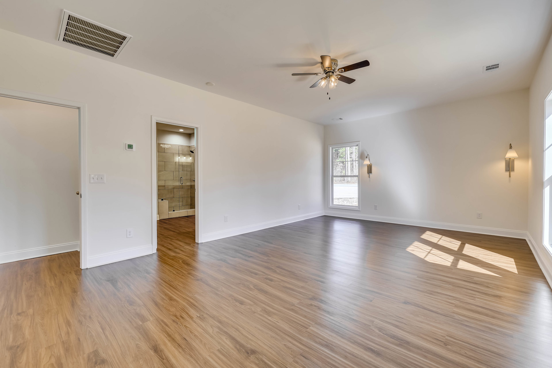 Wood flooring and white walls in a bright room featuring a ceiling fan with lights, a ceiling vent, a window overlooking trees, and a glass shower door with tiled wall.