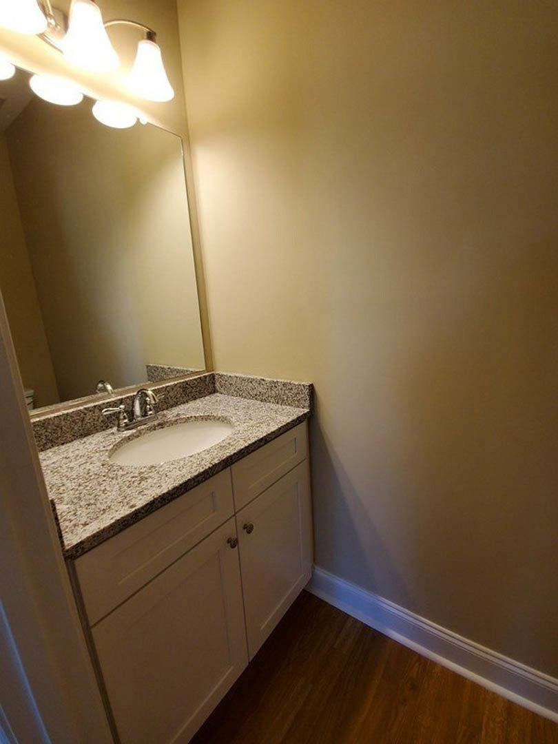 White ceramic sink with chrome faucet set in a wood vanity, rectangular mirror above, light fixture mounted on tiled wall, wood flooring visible