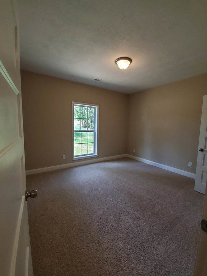 Neutral-toned carpeted room with white walls, large window overlooking green lawn, ceiling light fixture, and white door with brushed metal knob