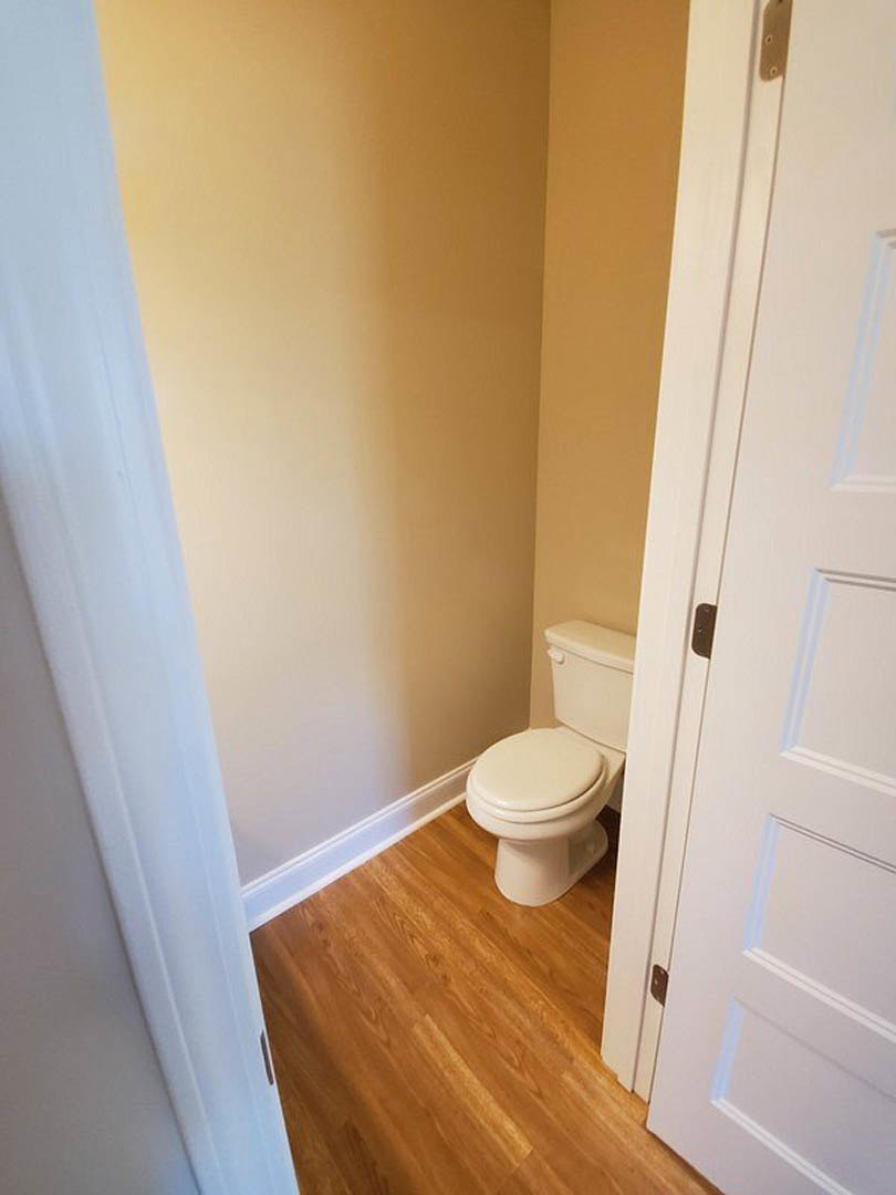 White toilet with closed lid on wood floor, white curtain and door partially visible, plaster walls and chrome door handle in bathroom.