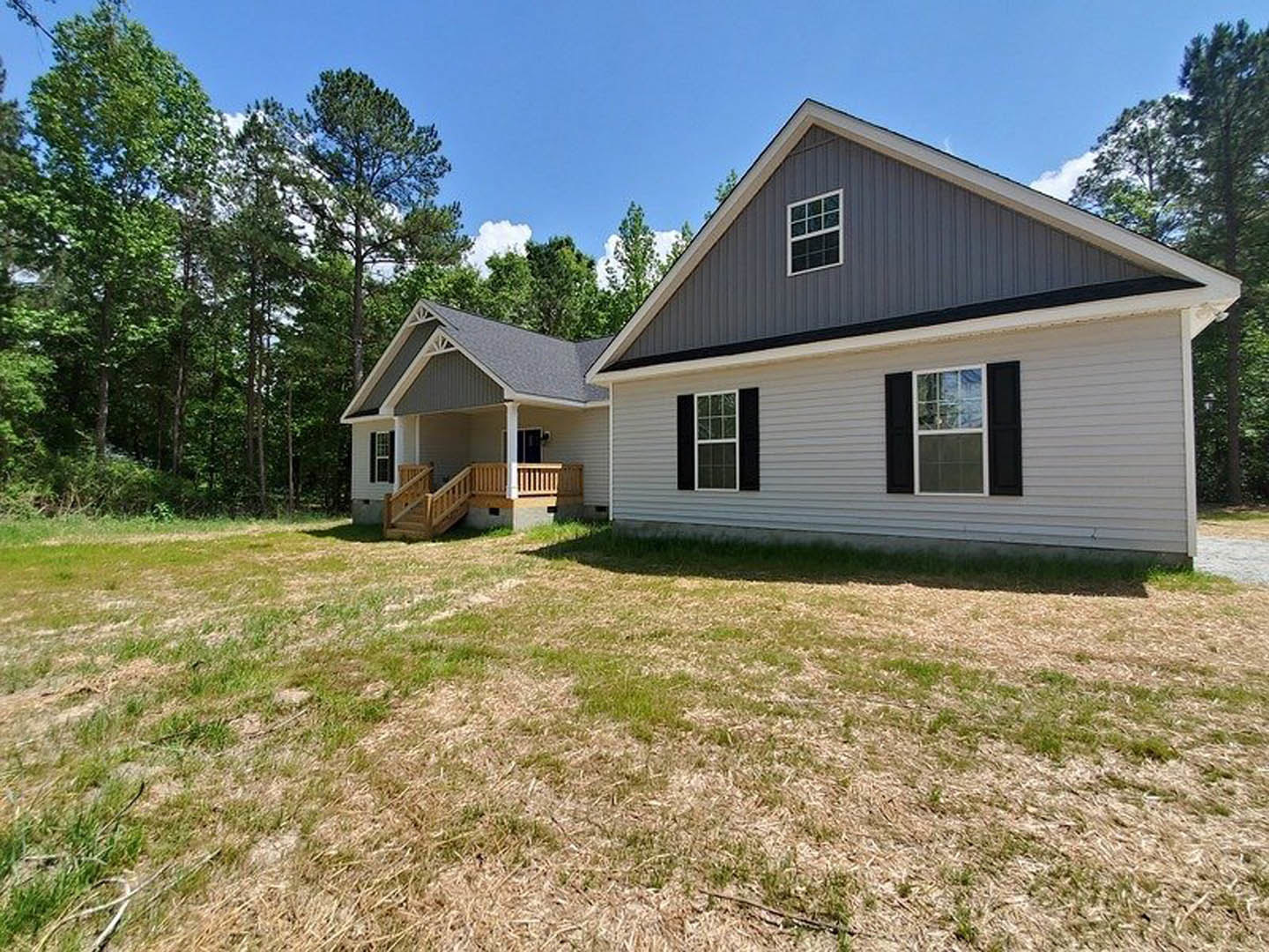Two-story house with black-shuttered windows, covered porch with wooden railing and stairs, surrounded by green grass lawn and mature trees in the background