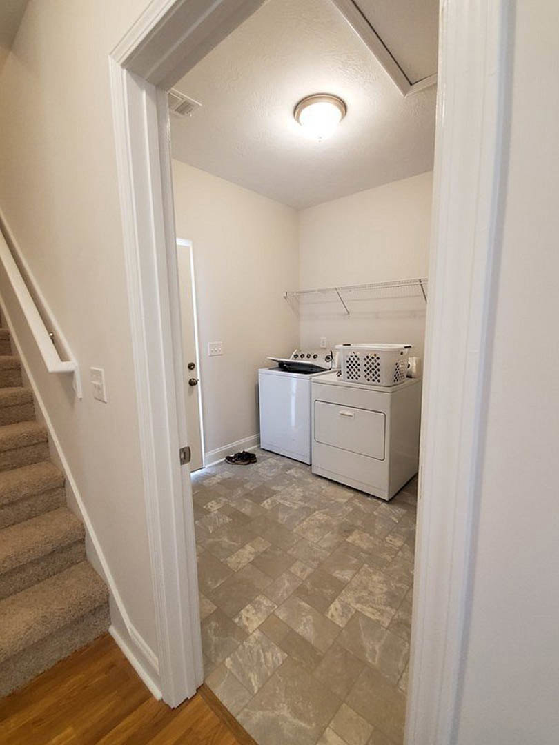 Laundry room with white washing machine topped by a basket and black item, white perforated container nearby, tile flooring transitioning to wood, staircase with wooden steps