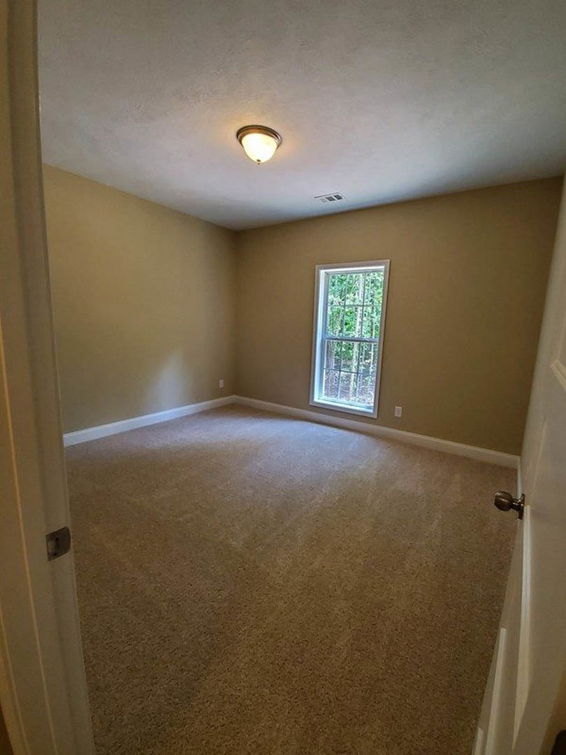 Beige carpeted room with white walls, large window showing leafy trees, ceiling-mounted light fixture, and door with silver knob
