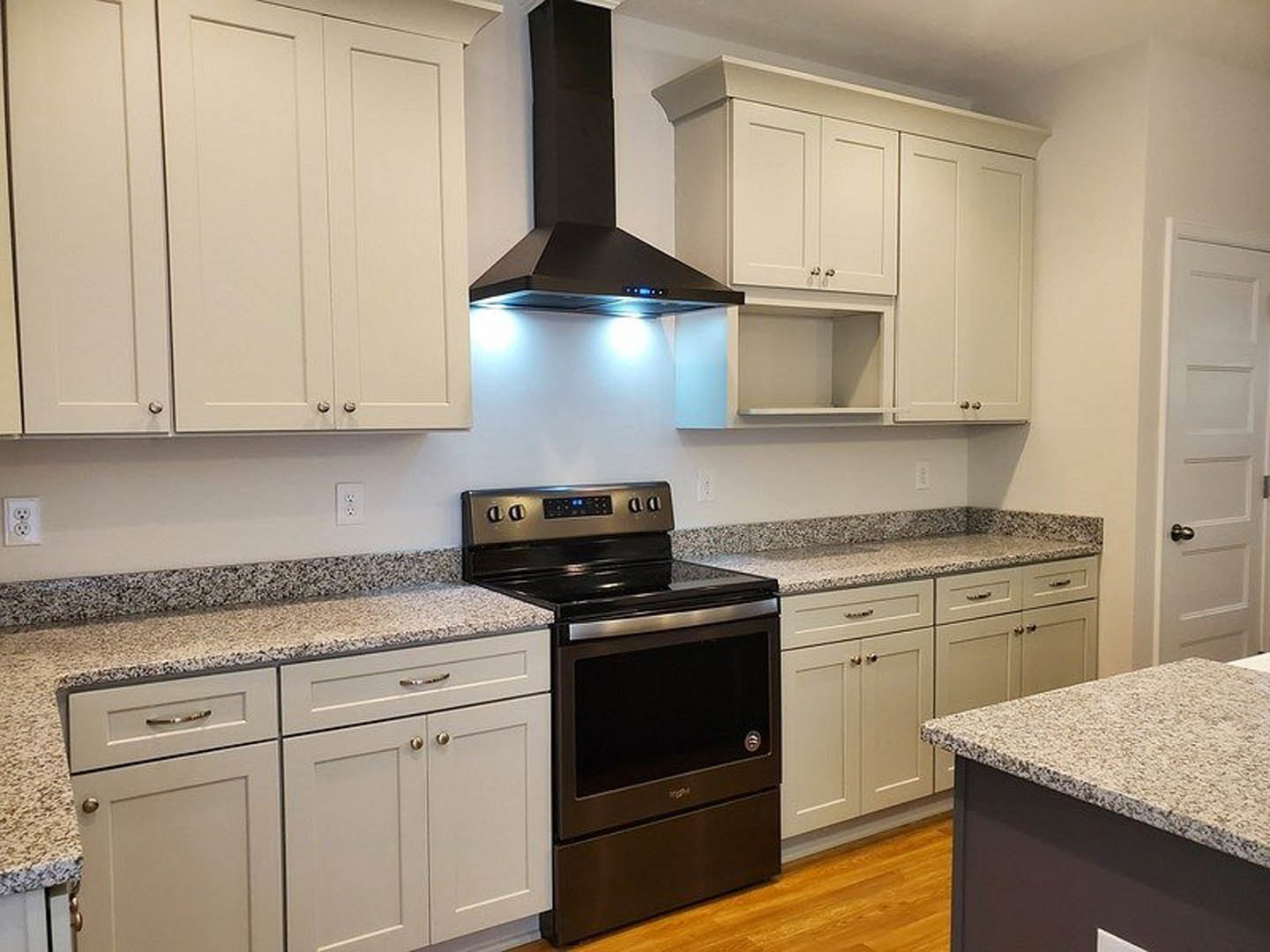 White kitchen cabinets with black stove and oven, wood flooring, black range hood with blue lights, white door with silver handle, light-colored countertop.