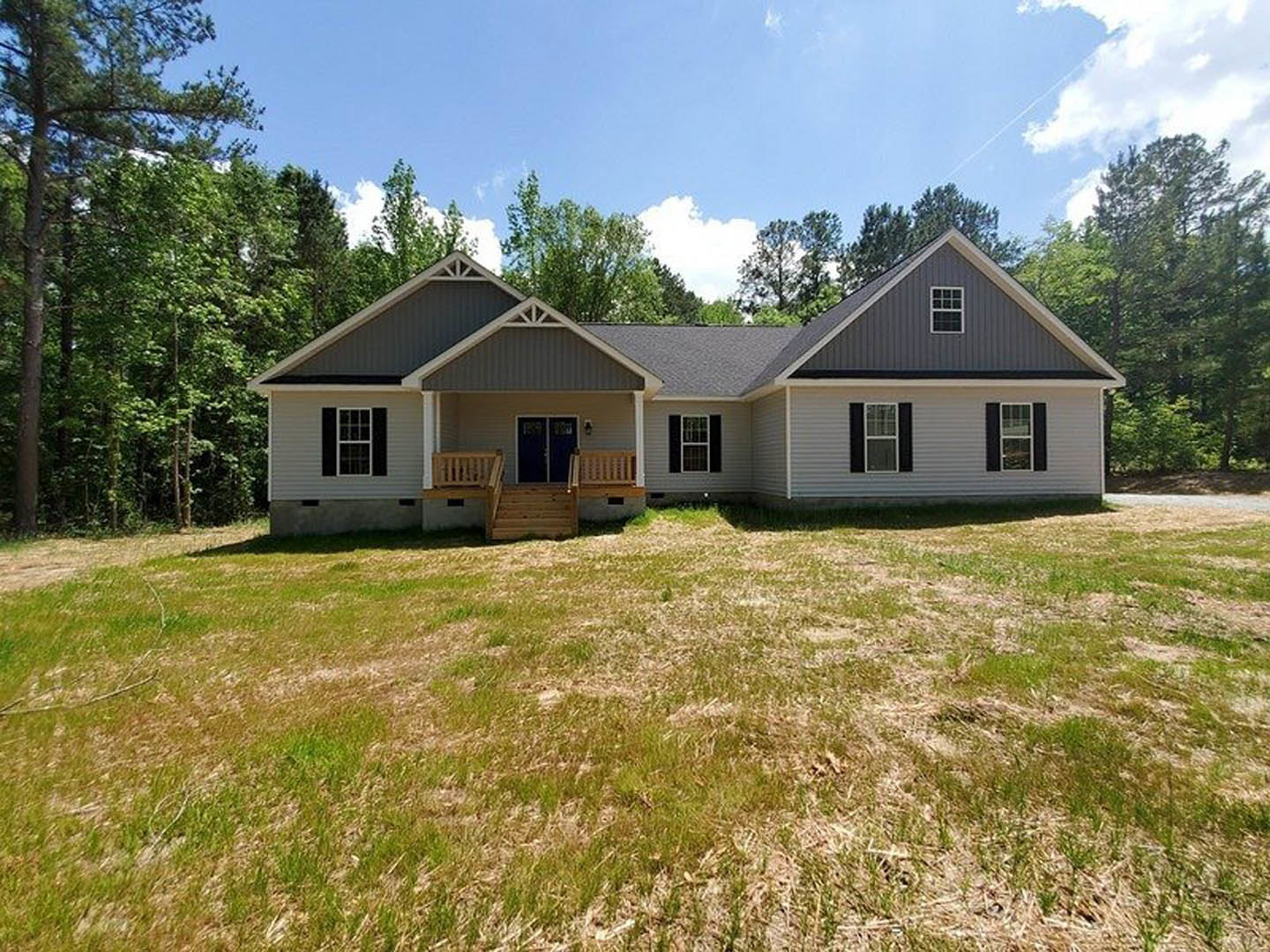 Two-story home with white-framed windows, wooden porch and stairs, expansive green lawn, wooden bench, and surrounding trees under a partly cloudy sky