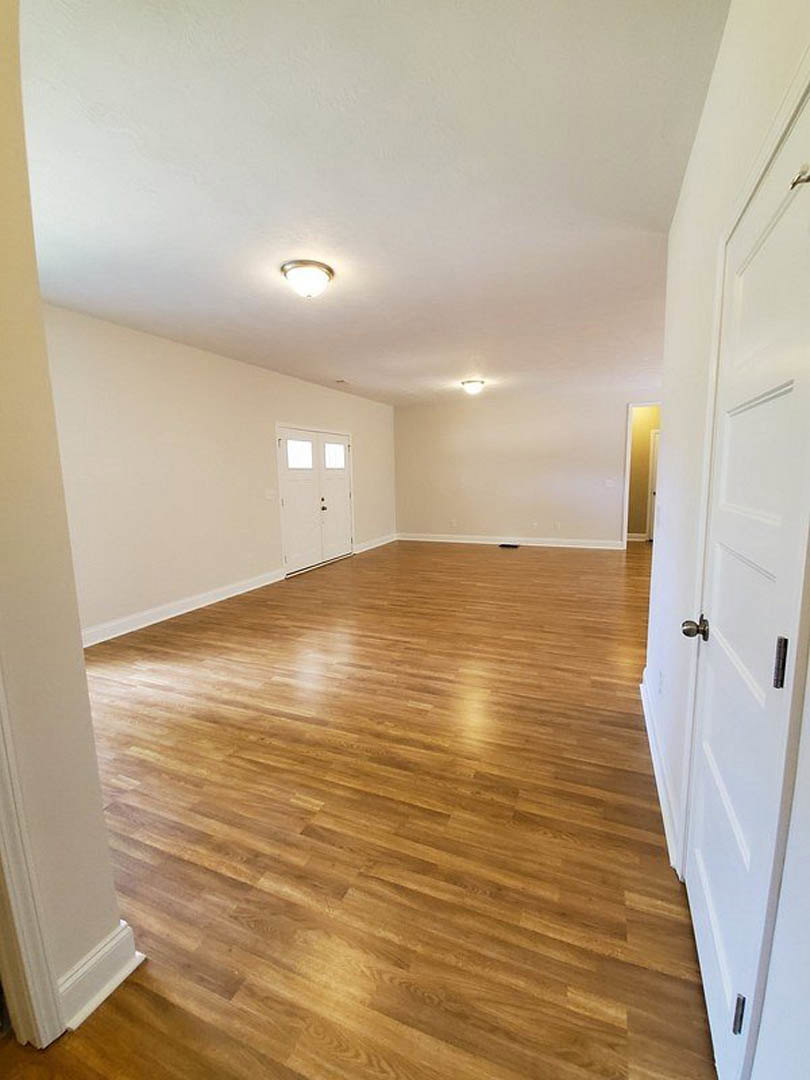 Spacious room with rich hardwood flooring, white walls, and a white door with glass panes; ceiling-mounted light fixture visible.