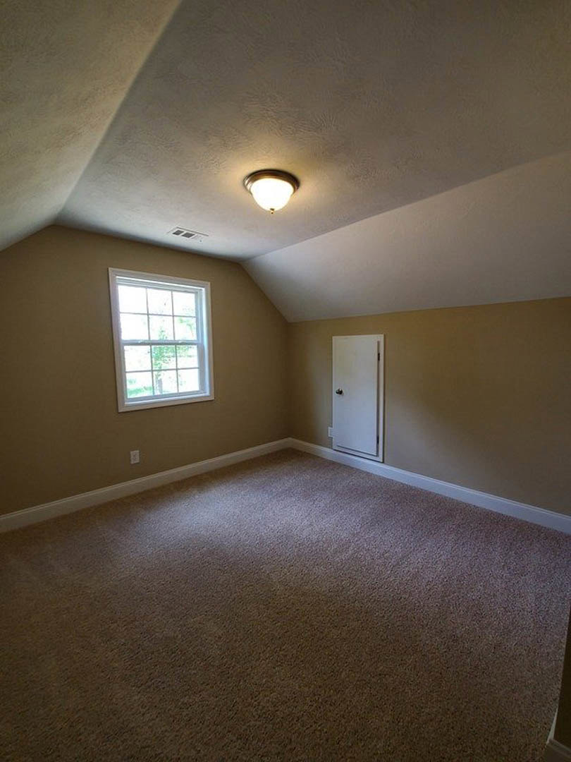 Carpeted room with white walls, multi-pane window, white door with silver handle, and ceiling-mounted light fixture