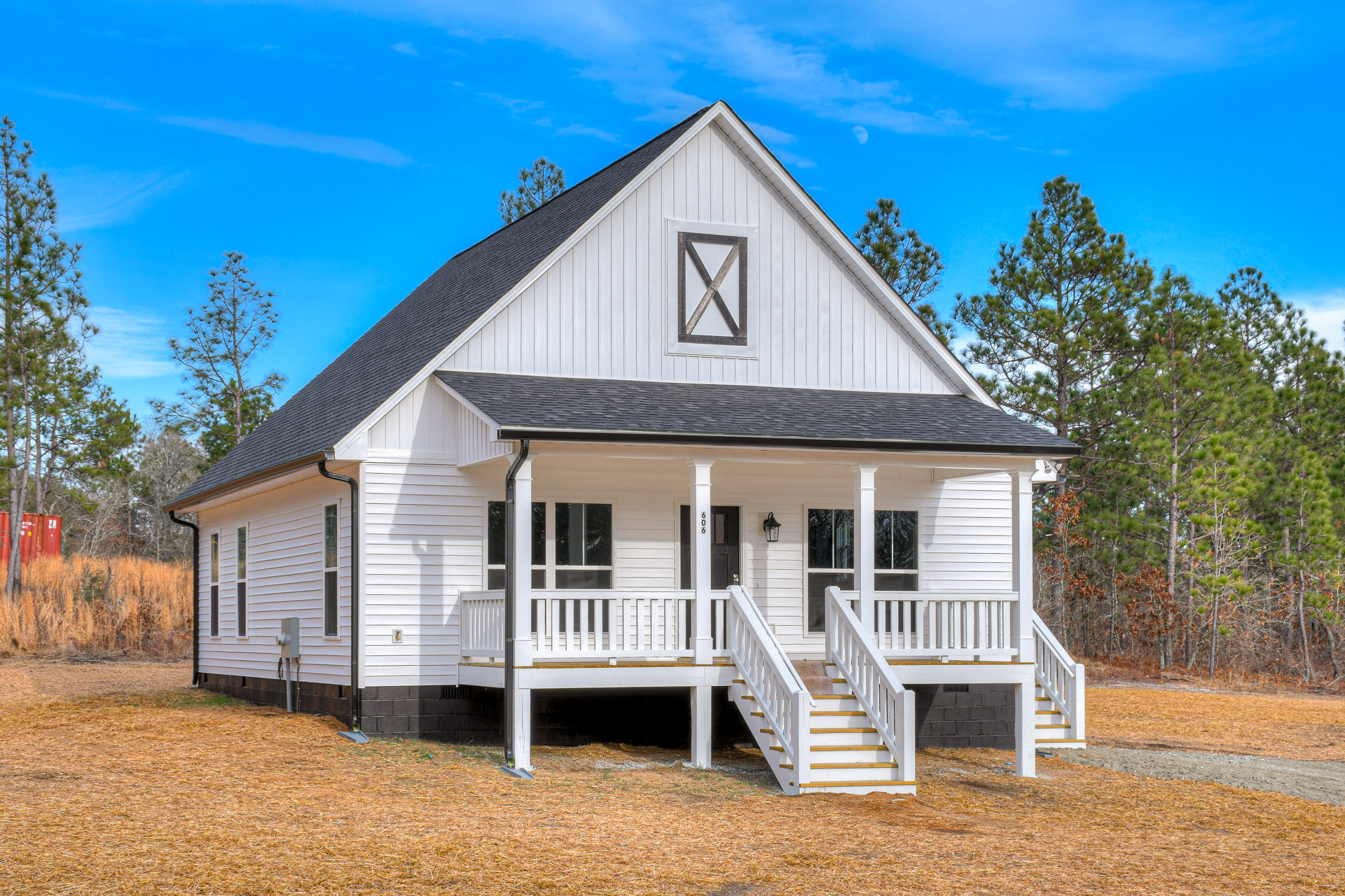 White siding house with black shingle roof, covered front porch with white columns and railings, exterior staircase, wooden porch framing with X detail, surrounded by tall grass