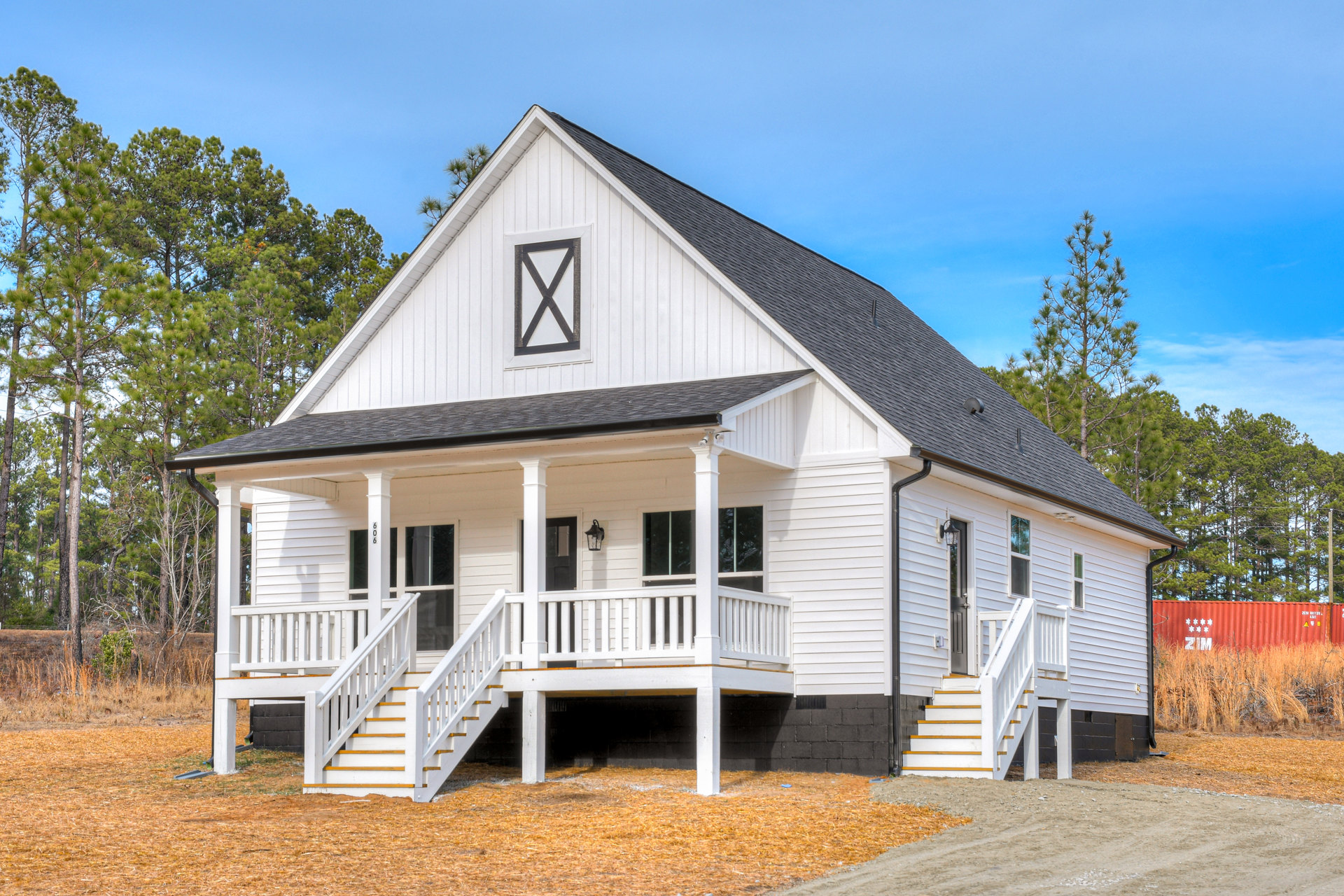 White siding house with black shingle roof, covered porch with white railings, exterior wooden stairs, red container with white stars and text on lawn, rectangular window with