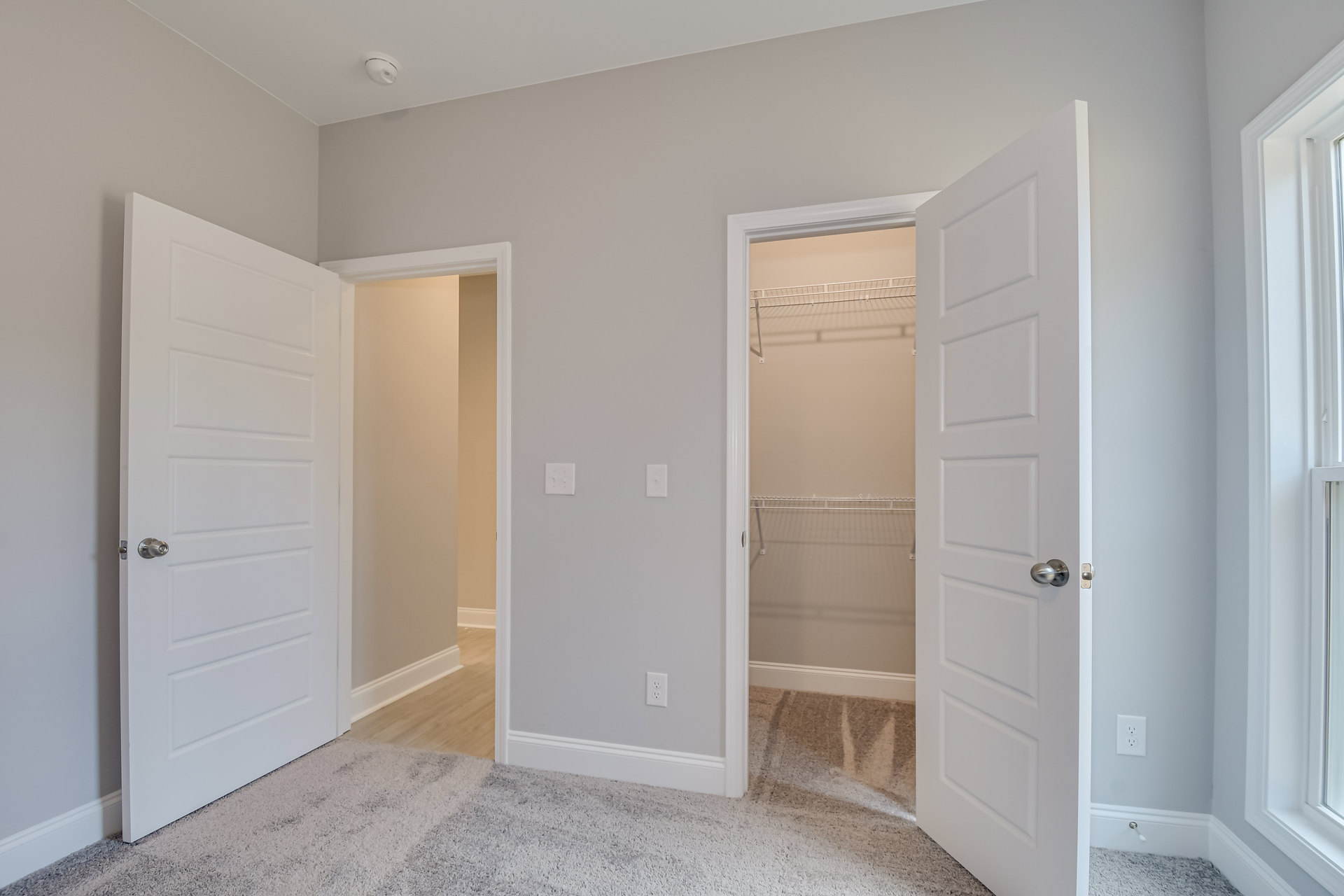 Open white closet door with silver knob reveals built-in white shelving, white carpet flooring, and light-colored walls with crown molding.