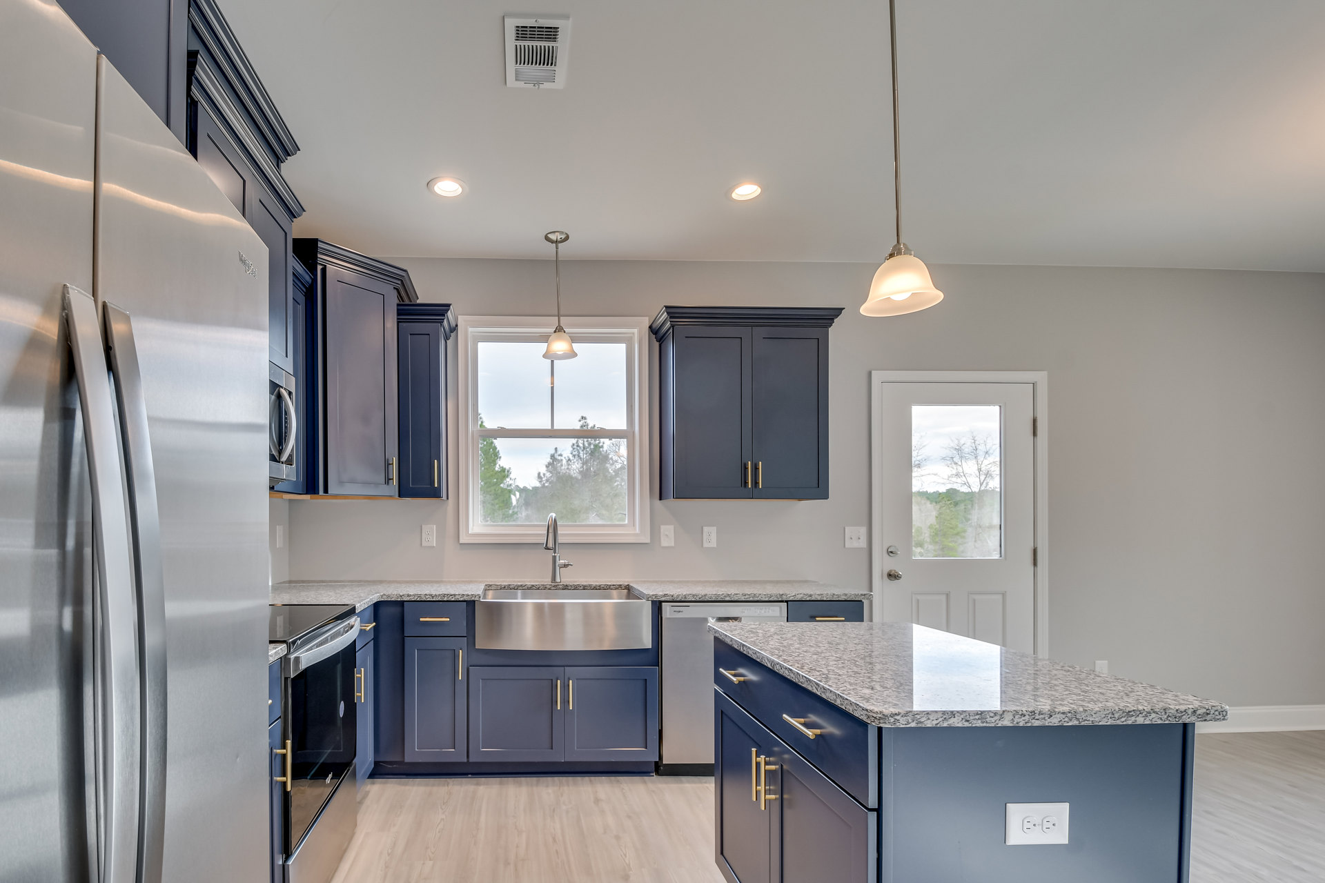 Blue kitchen cabinets with brushed metal hardware, granite countertops, stainless steel sink, window above with pendant light, white door with glass panel, and marble-topped