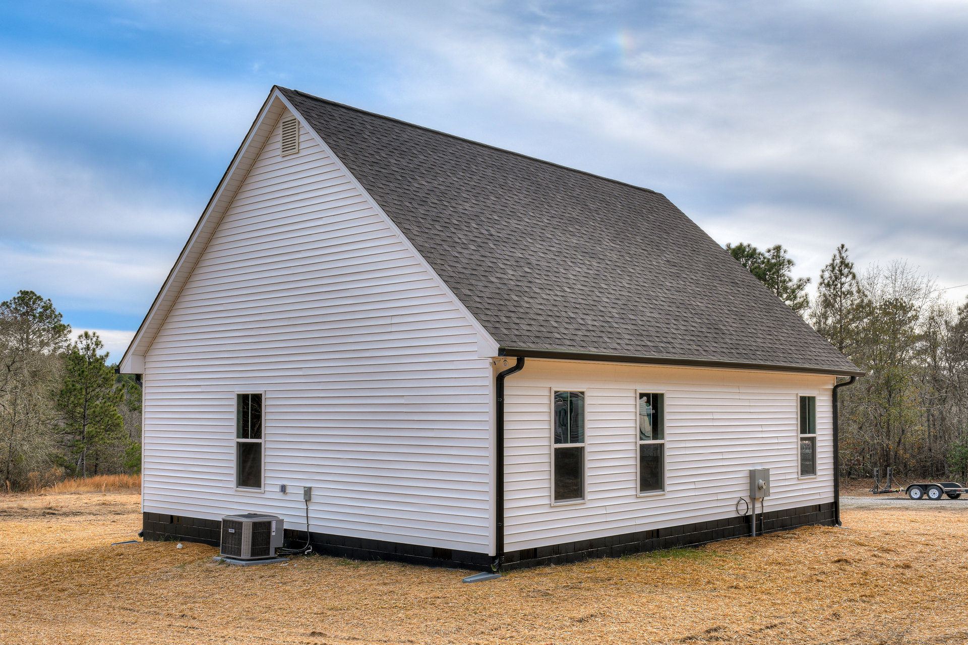White siding house with black shingle roof, multiple windows with white frames and curtains, large air conditioner unit on concrete pad, surrounded by trees under partly cloudy