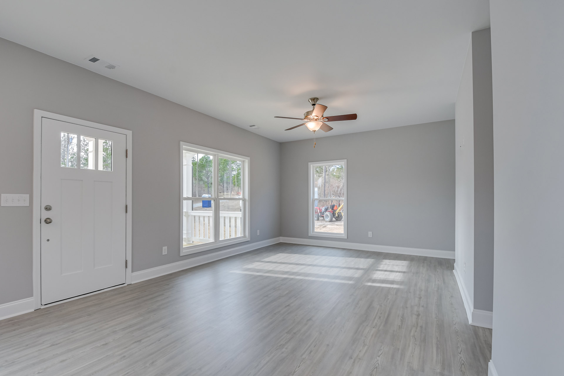 Wood-floored room featuring a ceiling fan with light, white door with window, multiple windows showing trees and a tractor outside, white window railings, plaster walls