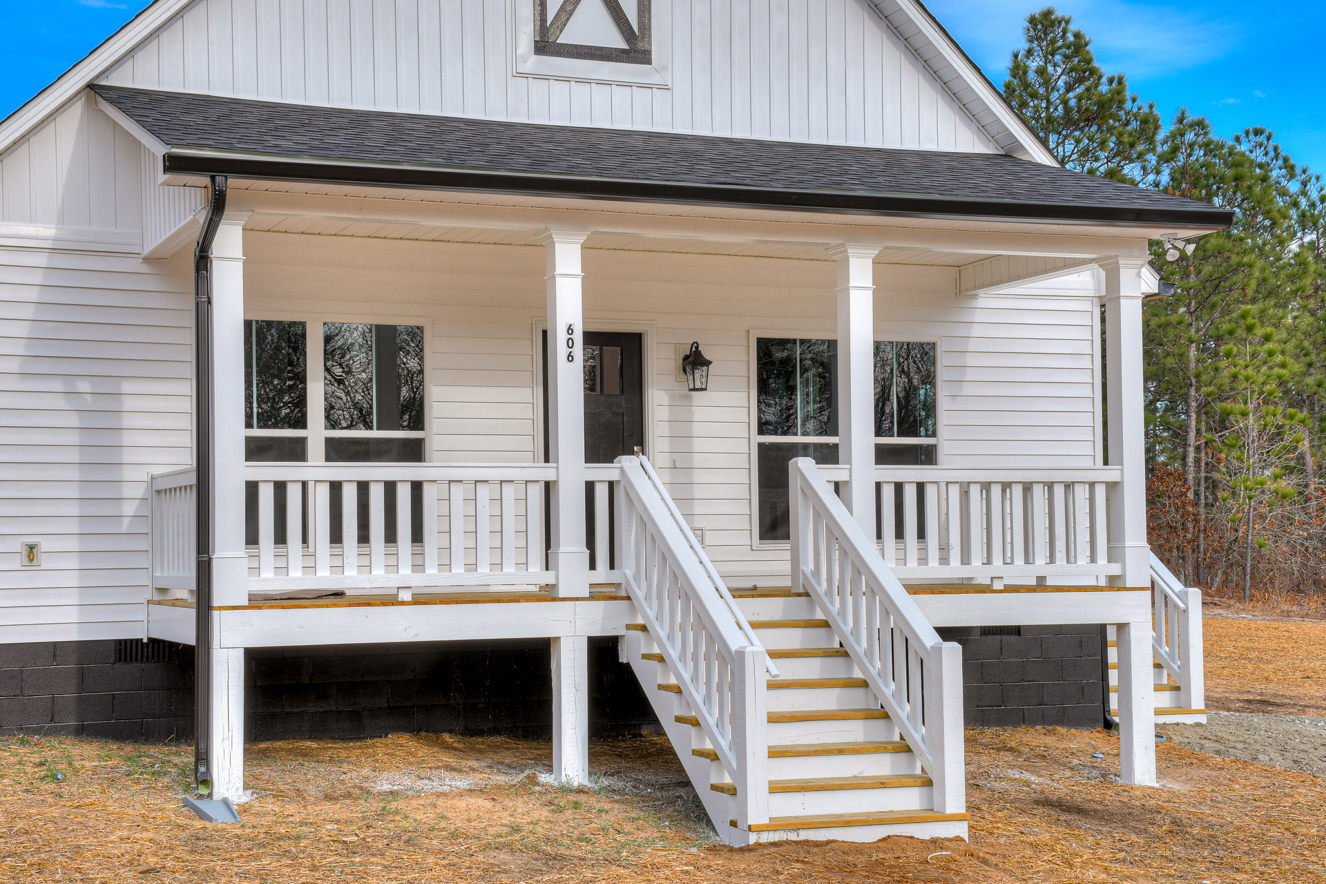 White wooden siding house with gabled roof, front porch supported by white posts, and outdoor stairs leading up to entry, surrounded by trees.