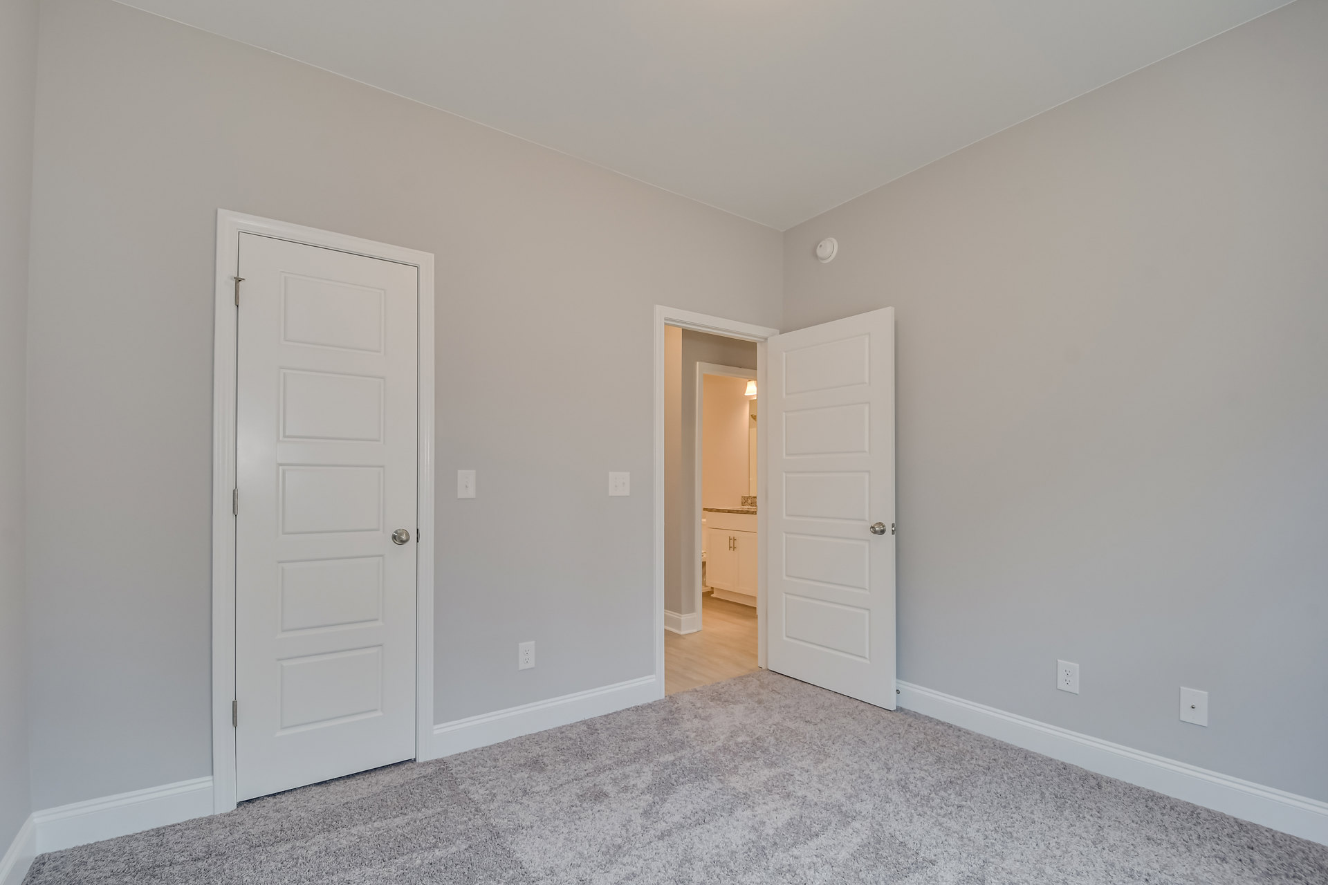 Bedroom with light carpet flooring, two white doors with silver knobs, one leading to a bathroom featuring a marble countertop