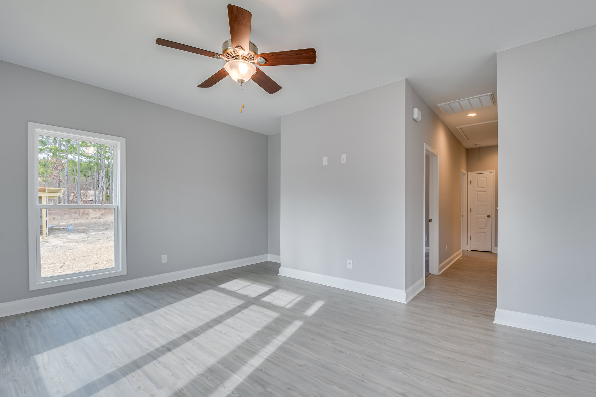 Ceiling fan with light fixture above wood floor, white door with silver knob, window showing trees outside, soft light illuminating neutral walls