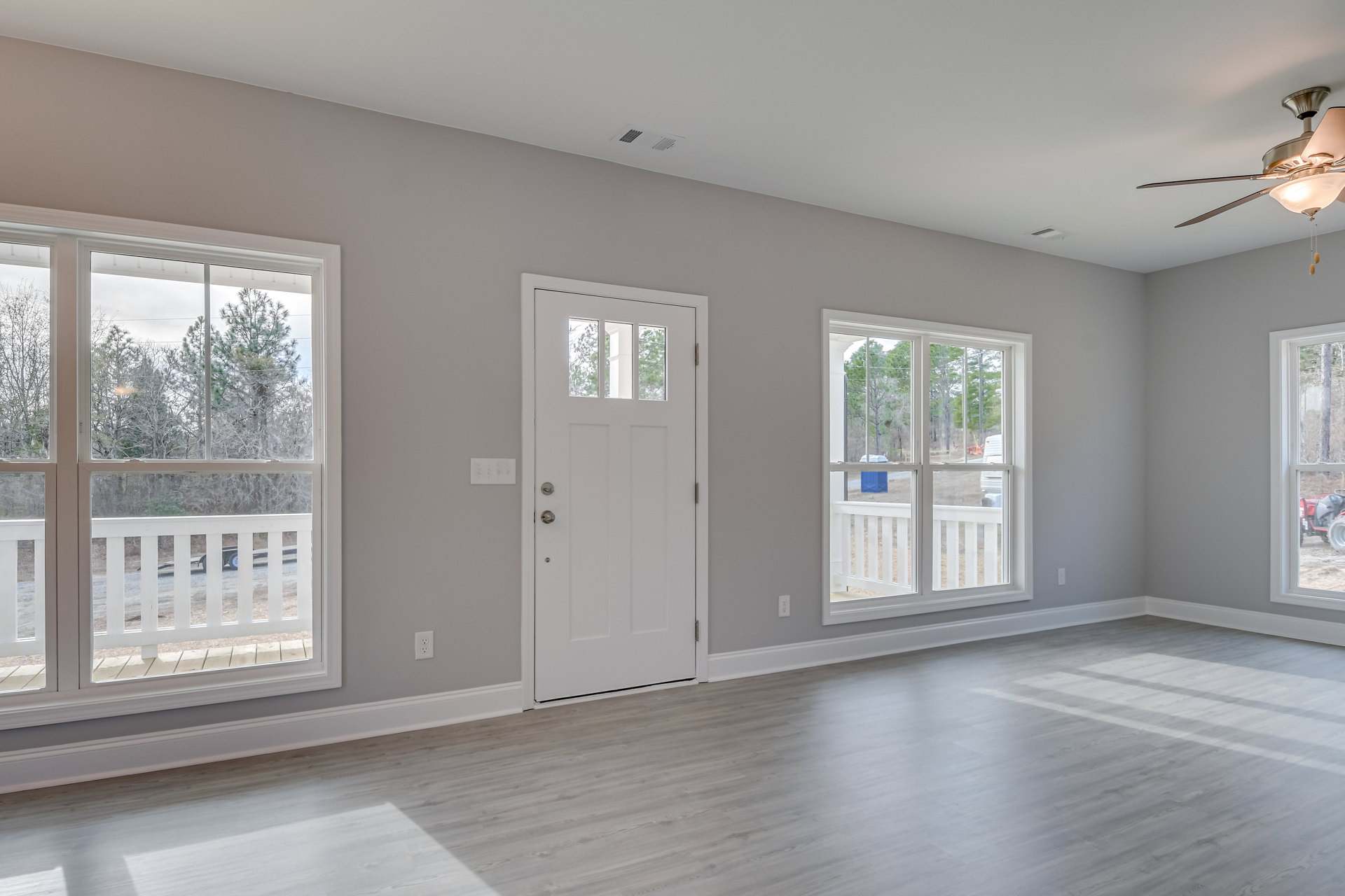 White paneled door with glass window, large windows showing trees outside, light gray walls, wood flooring, ceiling fan with light fixture