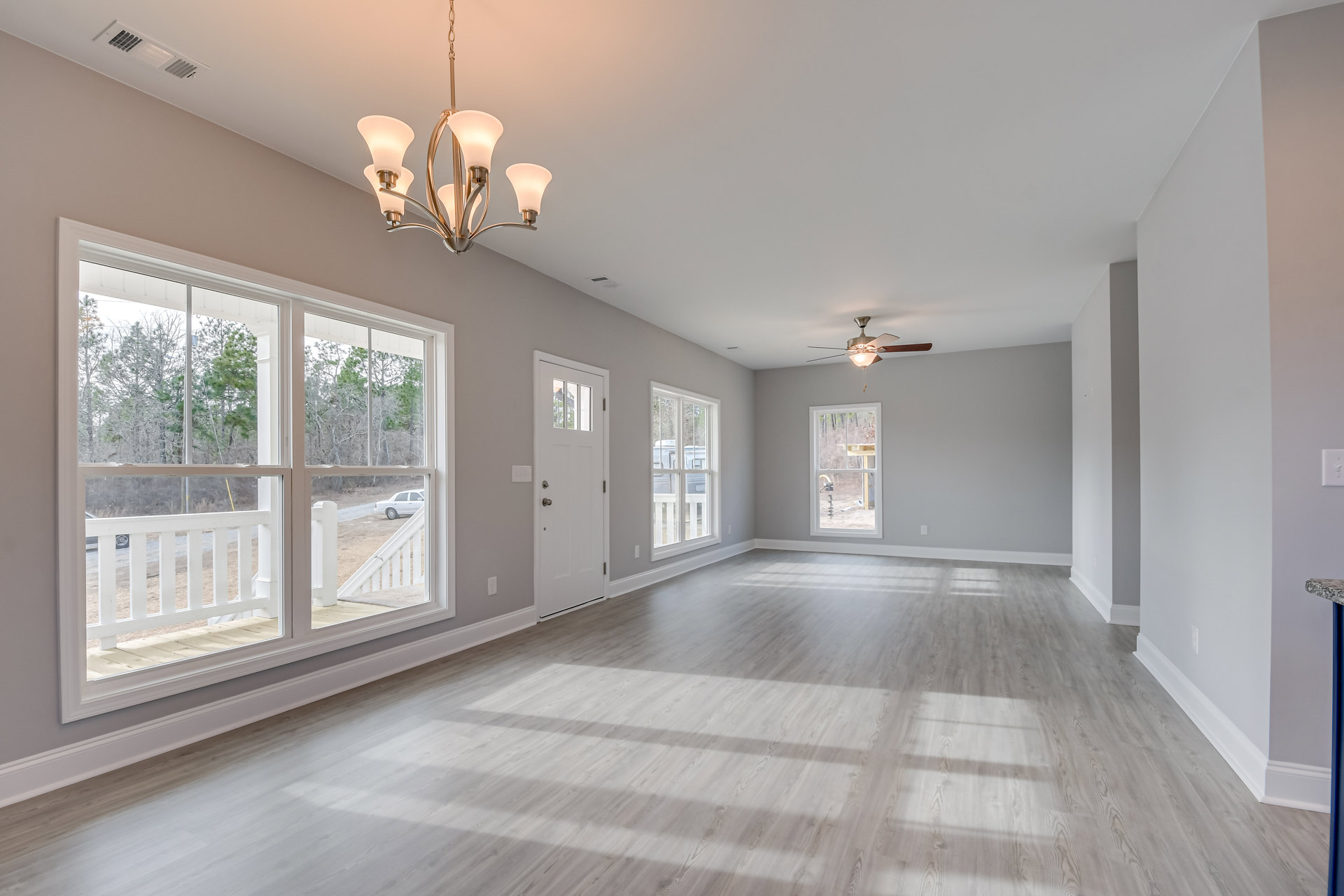 Chandelier hanging from white plaster ceiling, wood laminate flooring, large windows with white frames overlooking green yard, white door with silver doorknob, decorative wall