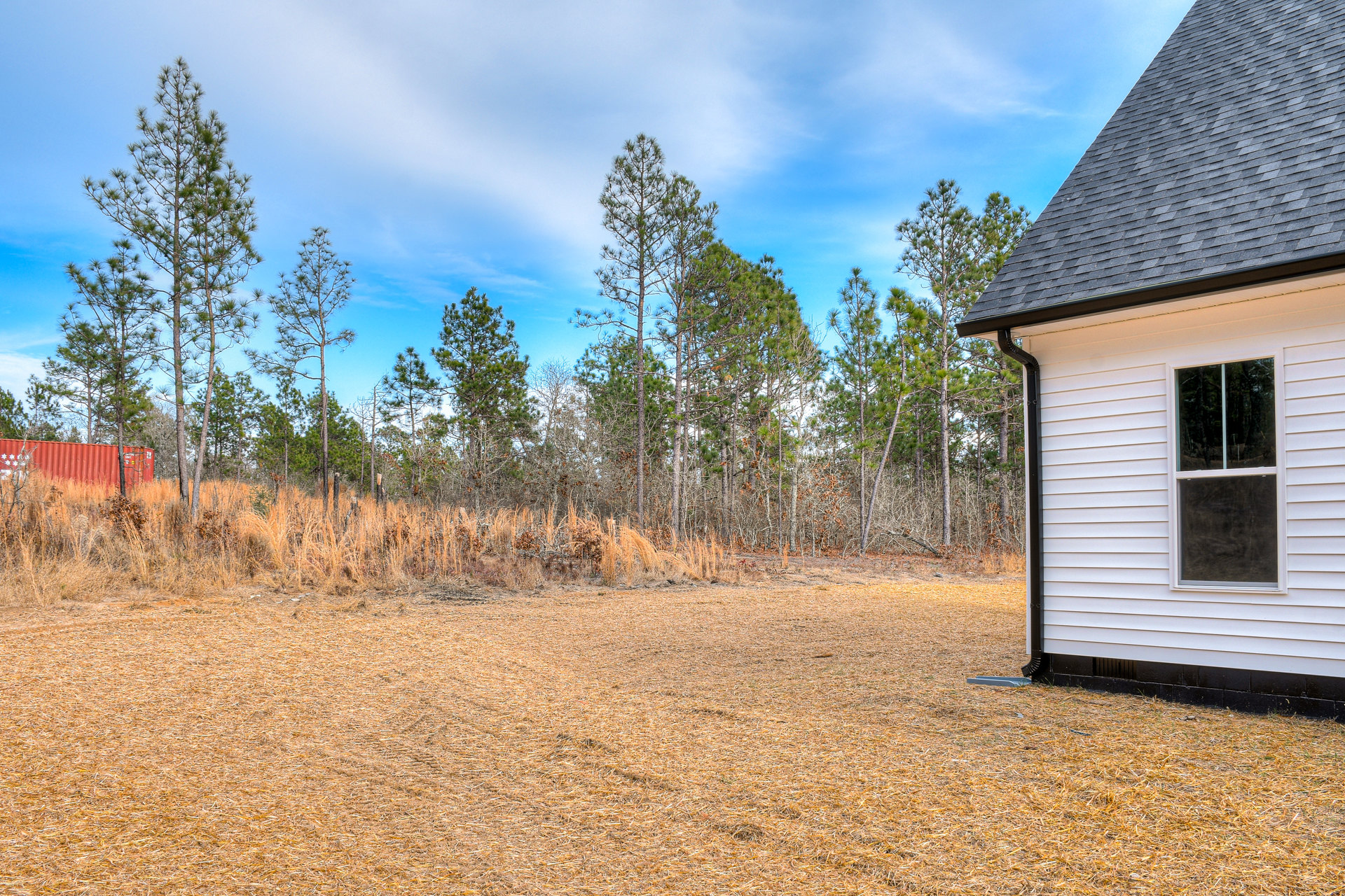 White cottage with black roof and white-framed windows, set in grassy field with scattered trees in background; red container sits in foreground.