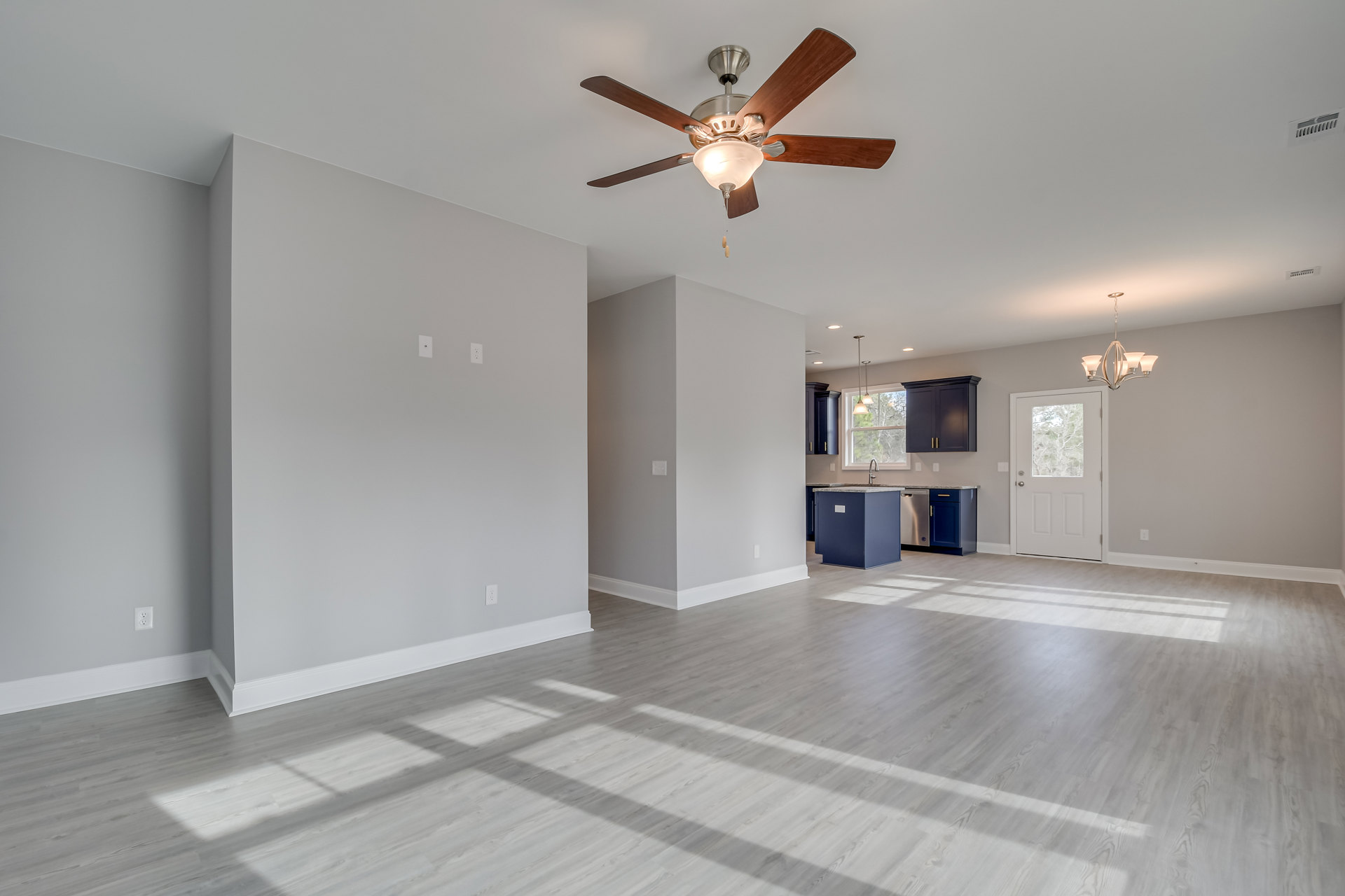 Open concept room with blue walls, white wood flooring, ceiling fan with light, blue kitchen island featuring a sink, white door with window, and white wall with electrical