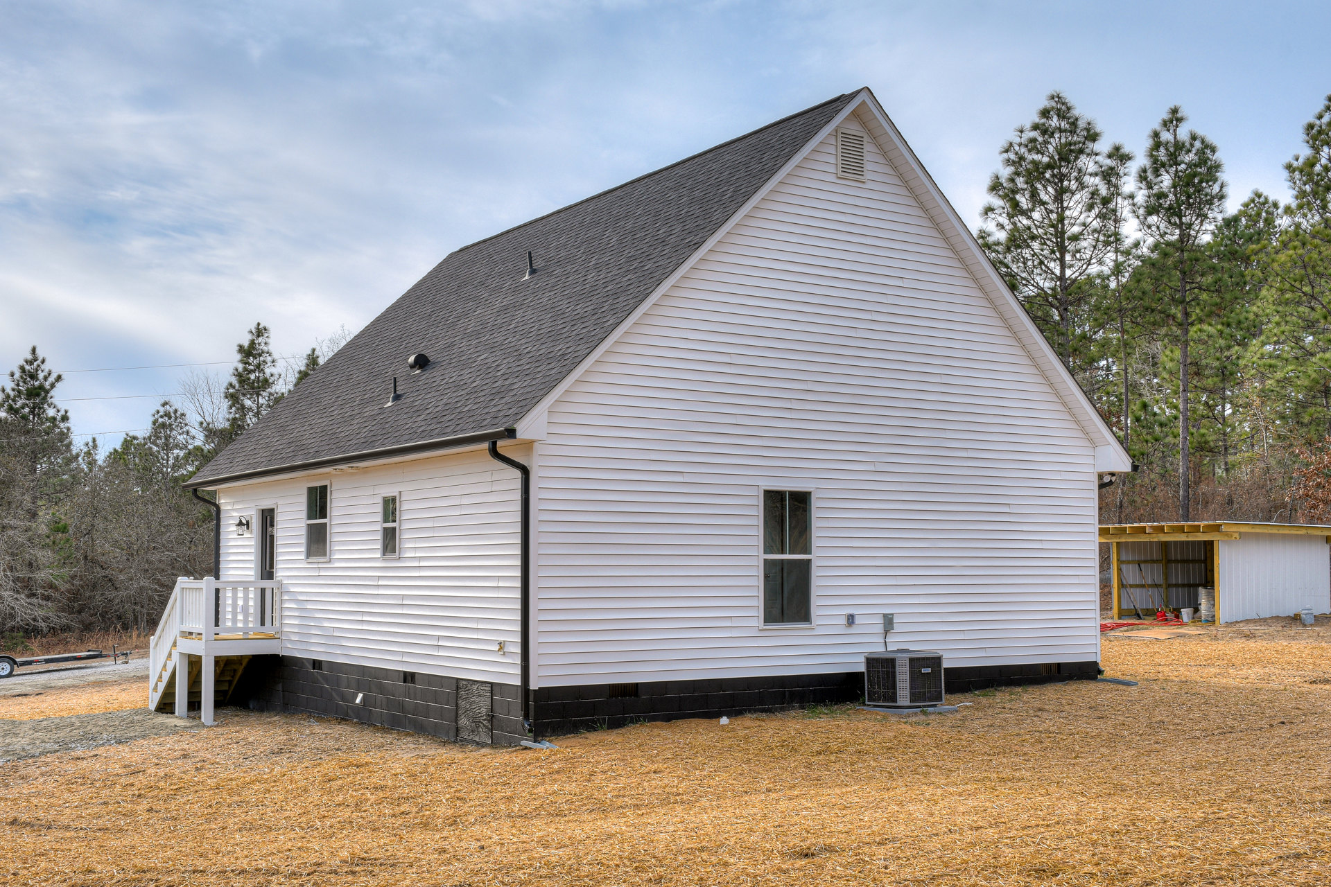 White siding house with black roof and trim, white front stairs, metal-roofed shed, visible window and outdoor air conditioner unit, surrounded by trees and sky.