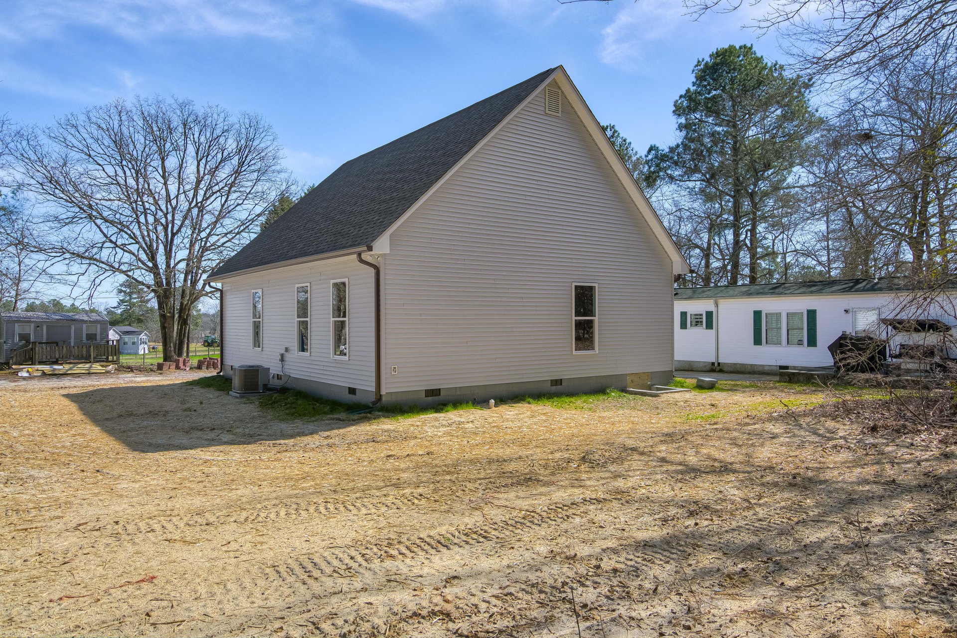 Gravel driveway leading to a farmhouse-style home with gabled roof, large windows, and a prominent air conditioner unit on a stand; mature tree with many branches in front, open