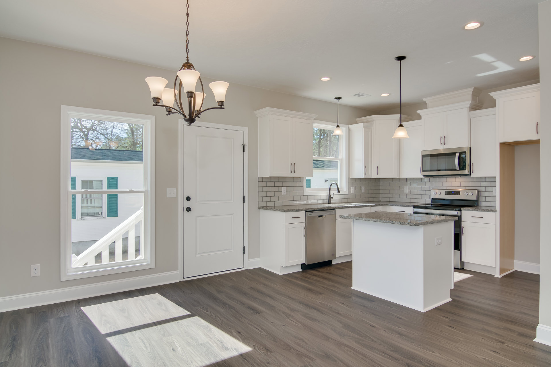 Kitchen with light wood flooring, central island featuring built-in stove, white cabinetry, stainless steel microwave, chandelier overhead, window with green shutters, white door