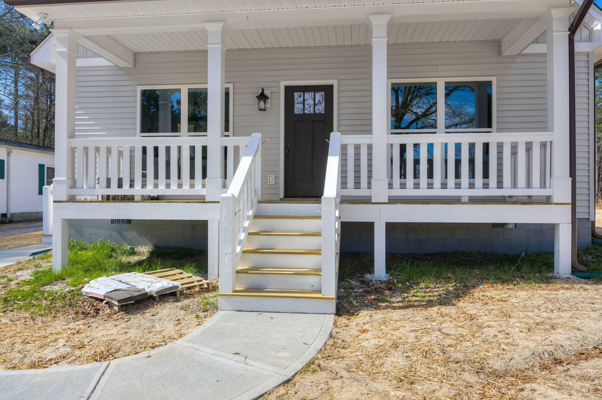 White house exterior with wooden porch, black glass-paneled front door, white railing, and stairs leading up to entry; green plants border the walkway.
