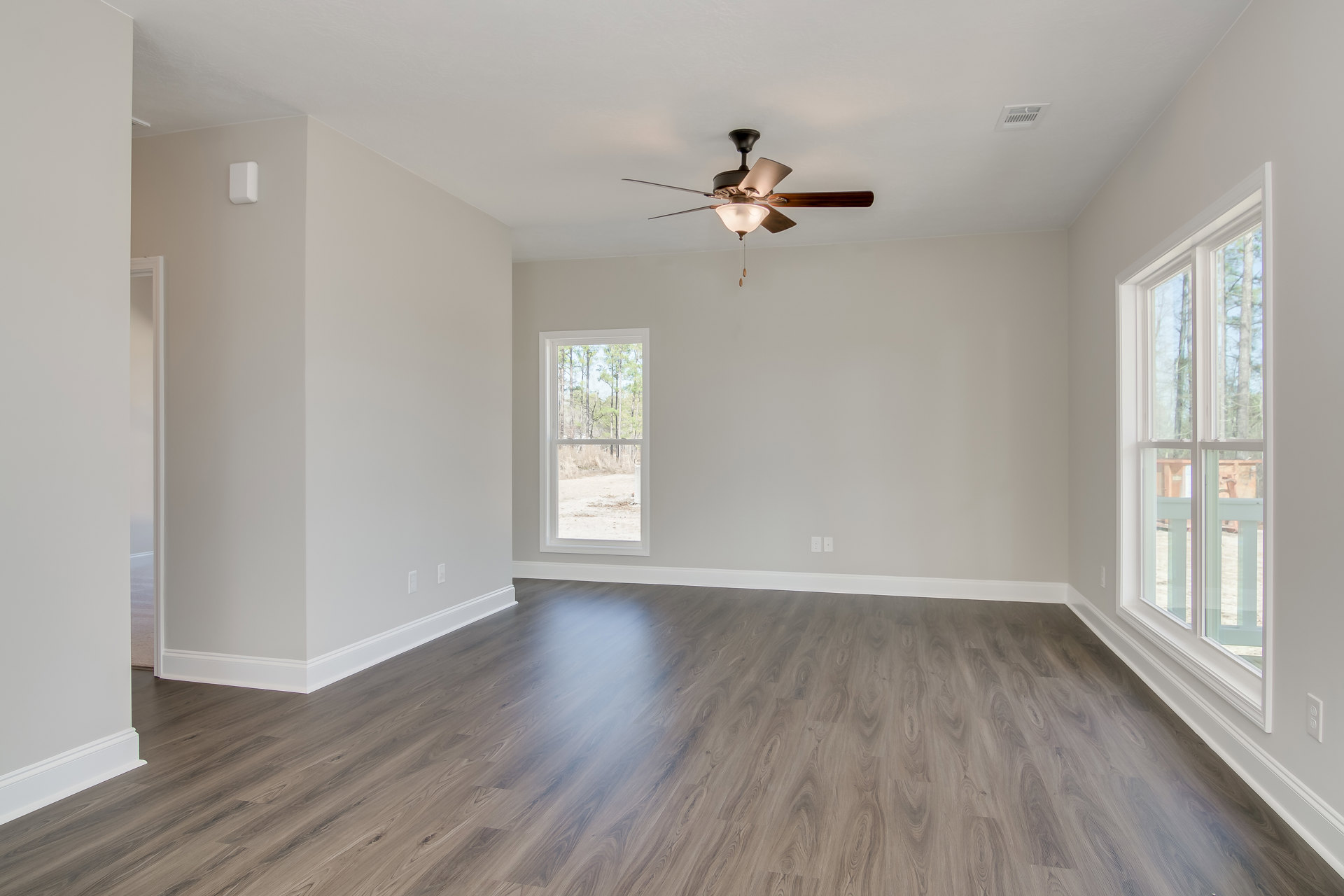 Ceiling fan with light fixture above hardwood floor, white plaster walls, large window revealing green trees outside