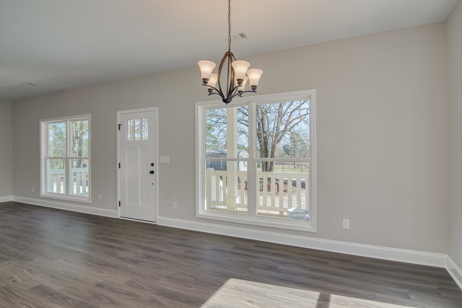 Five-light chandelier hanging from white ceiling, wood laminate flooring, white door with black knobs, large window with view of green trees, white walls with decorative molding.