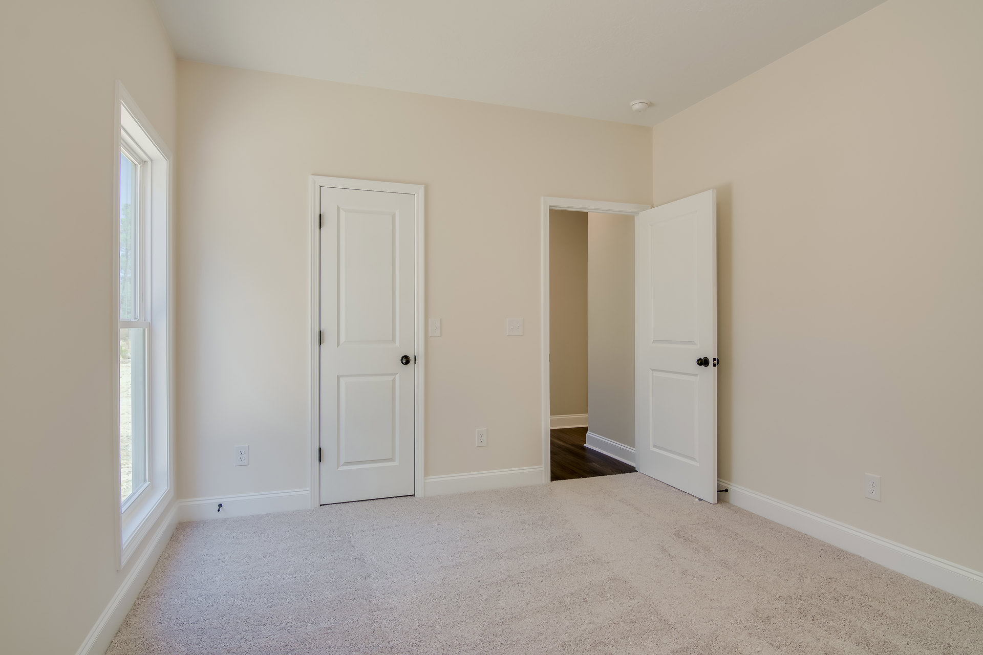 Carpeted room with two open white doors, white framed window, white walls, black door knobs, and simple molding.
