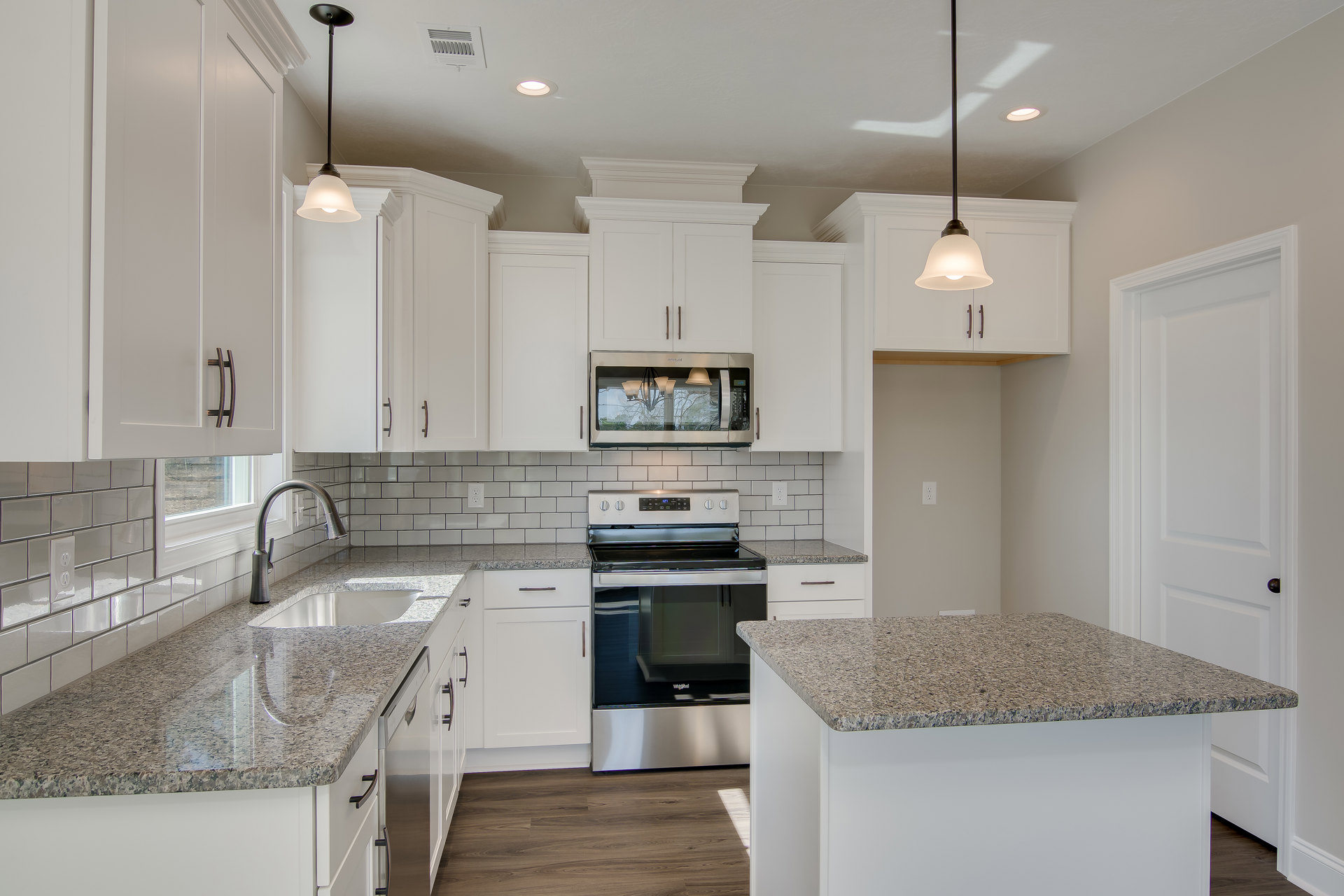 Granite countertops, white shaker cabinets, stainless steel oven with glass door, built-in microwave, modern light fixture, white walls, black door handle, corner support pole