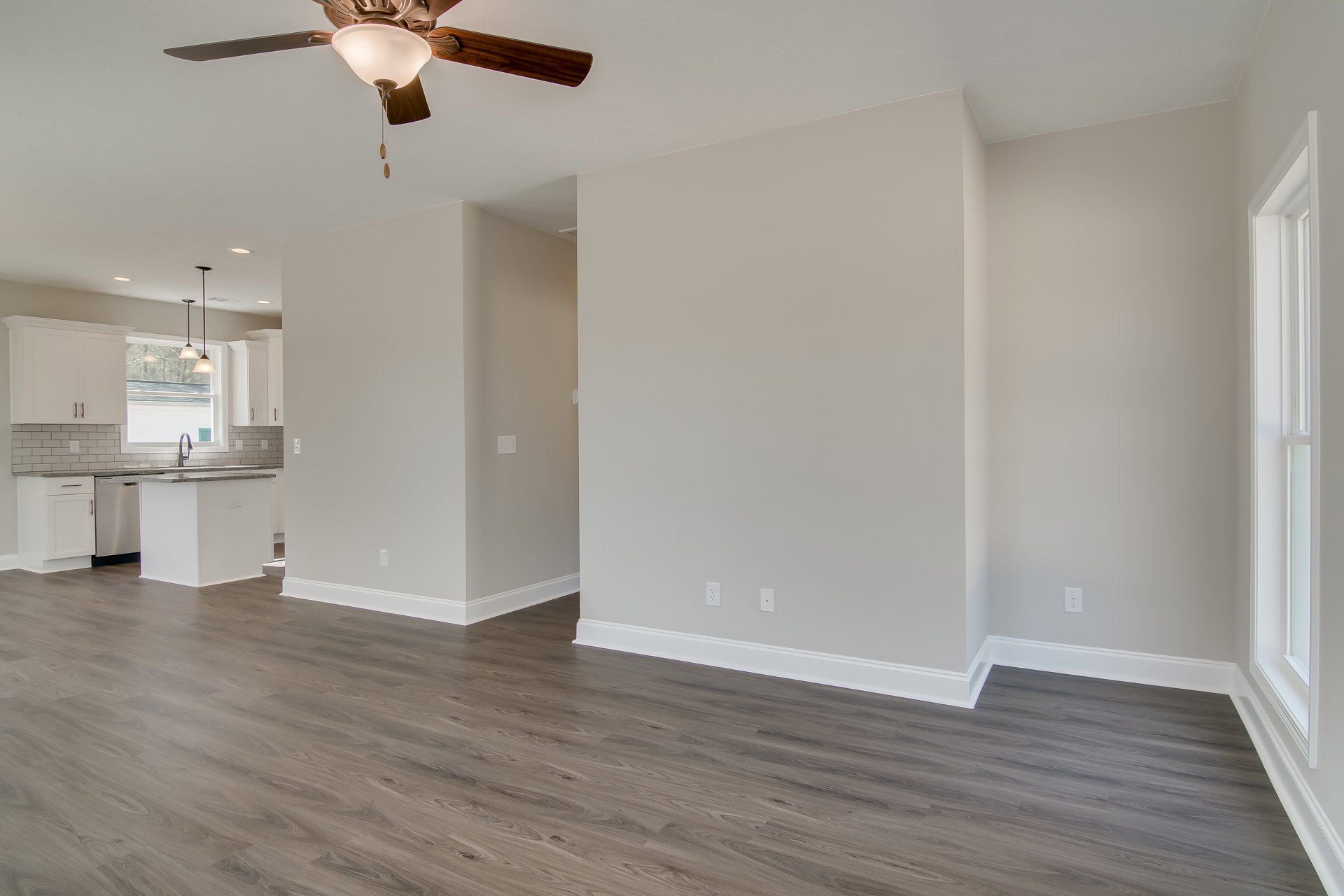 Ceiling fan with light fixture above wood floor, white walls, and white kitchen island with marble countertop; kitchen sink beneath window in open-plan room