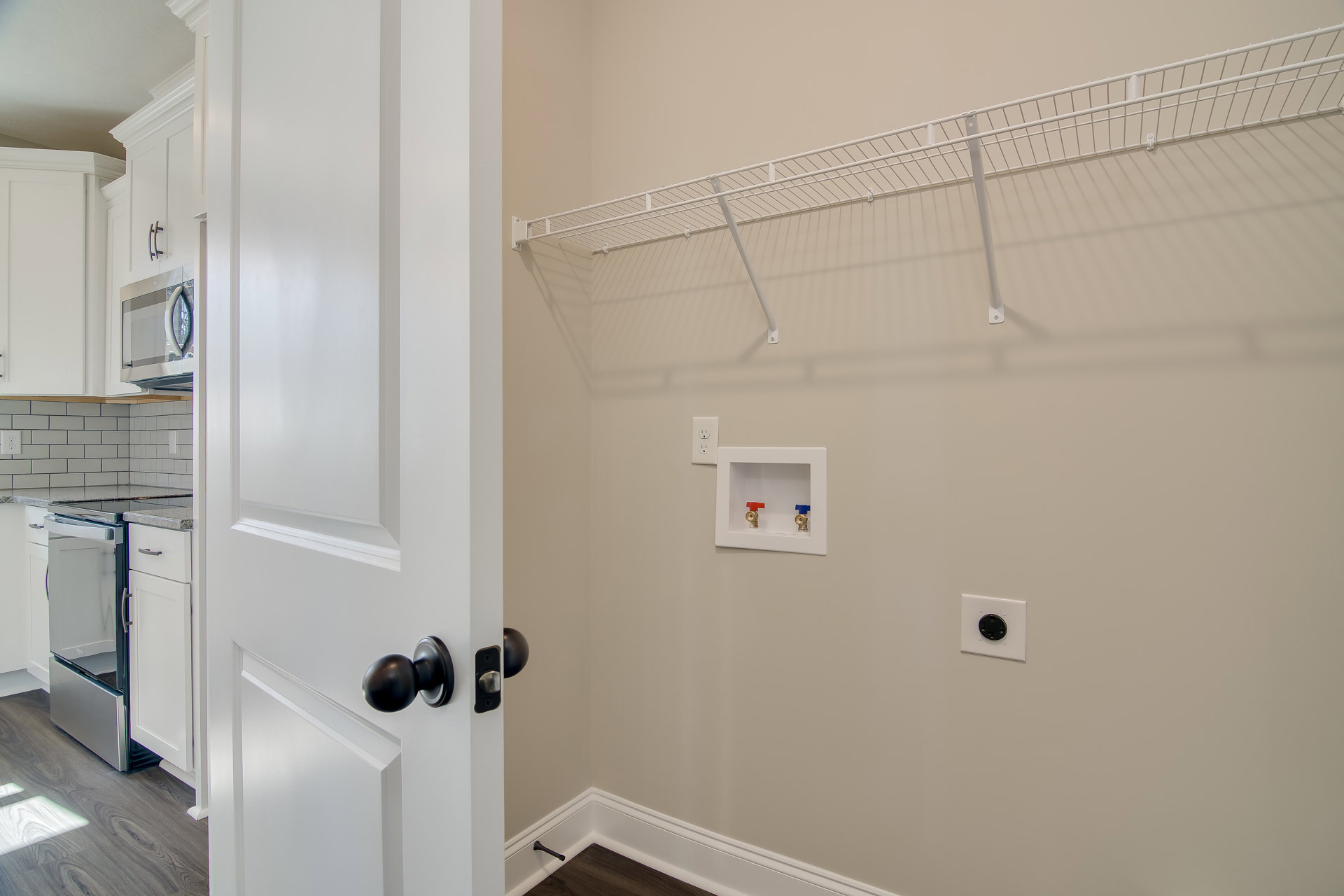 White door open to a closet with white shelving, visible plumbing valves, electrical socket, refrigerator, and white cabinets on wood flooring.