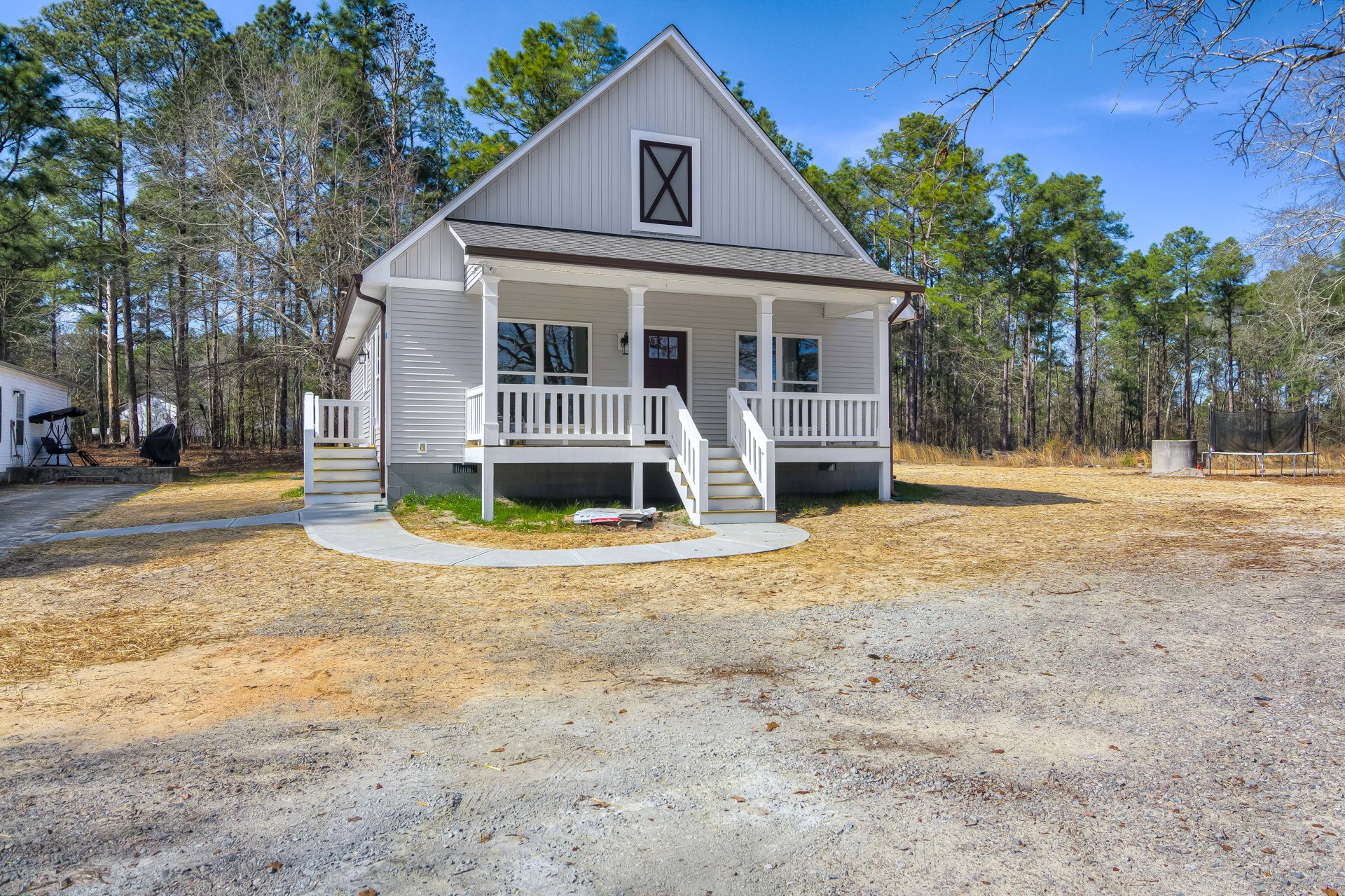 White house with covered porch, white railings, gravel driveway, and mature trees in the background