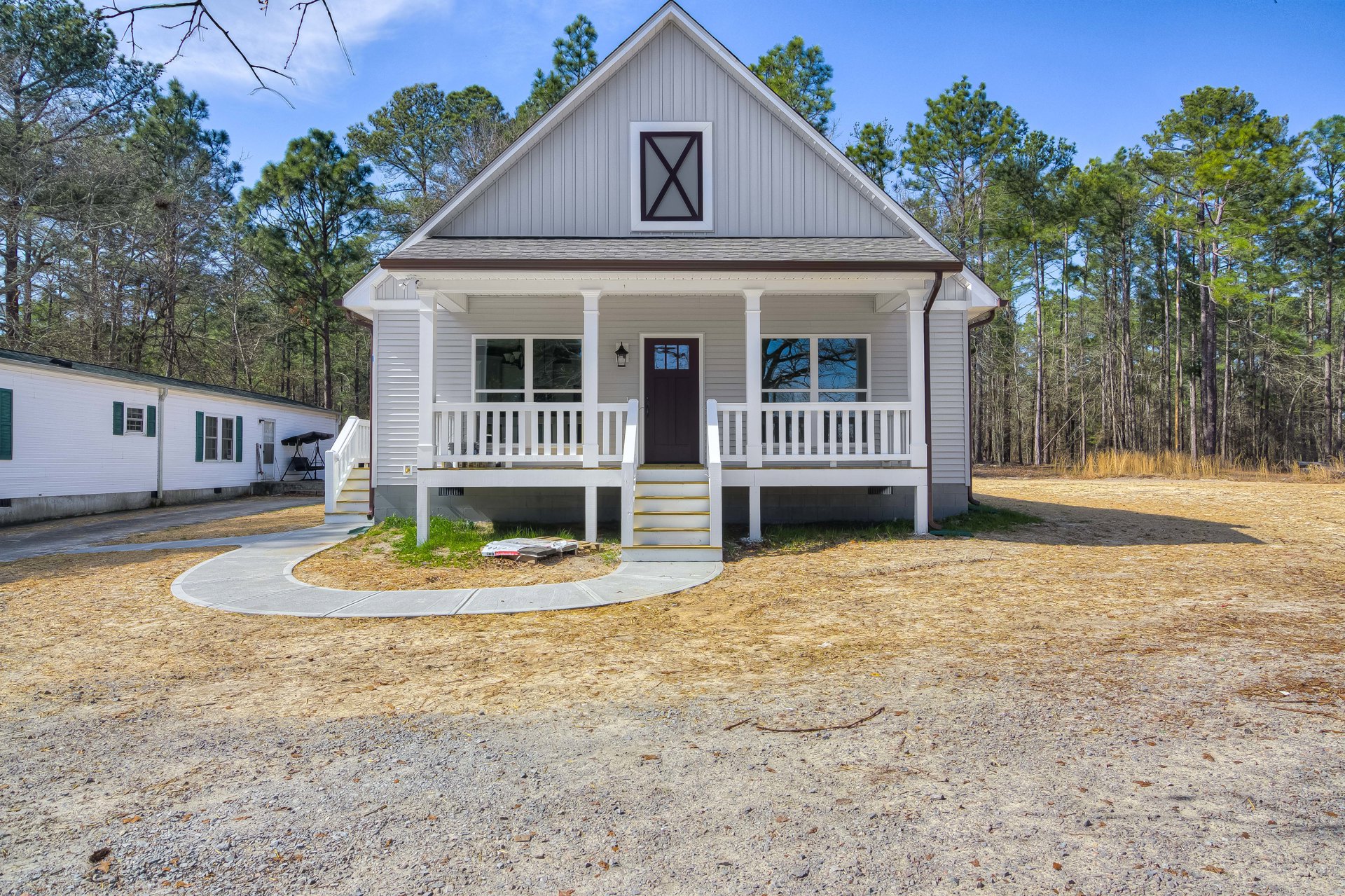 Two-story home with gray siding, black front door, white porch railing, concrete driveway, and mature trees in the background