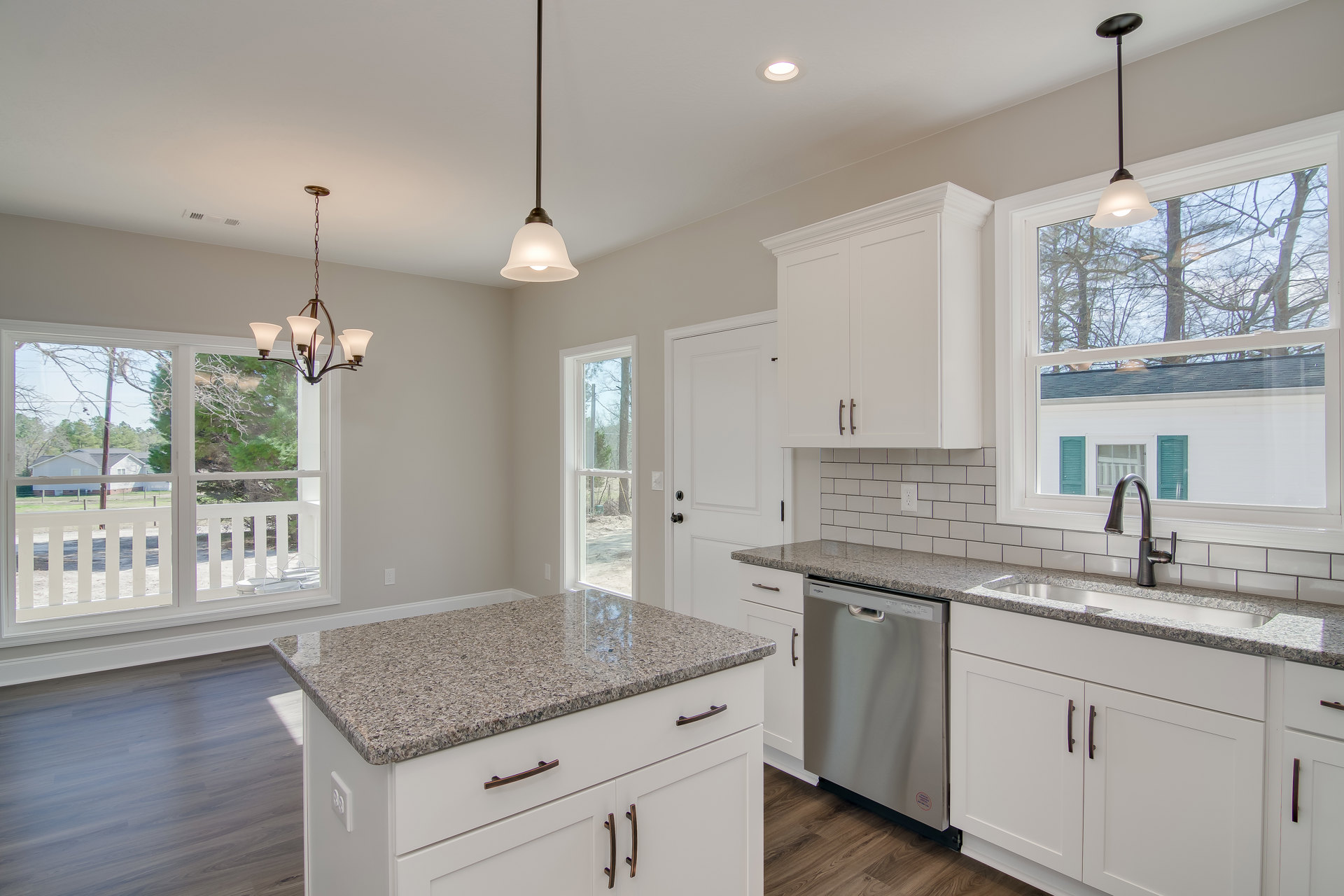 White kitchen cabinets with granite countertops, stainless steel dishwasher, window above sink, chandelier with white shades, black support pole, and light-colored walls.