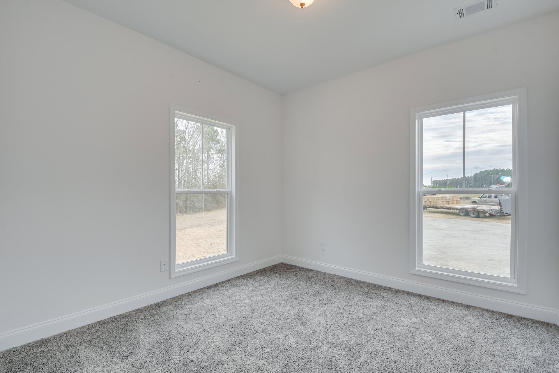 Carpeted room with multiple windows, views of trees, grassy field, road, and parked truck with trailer outside