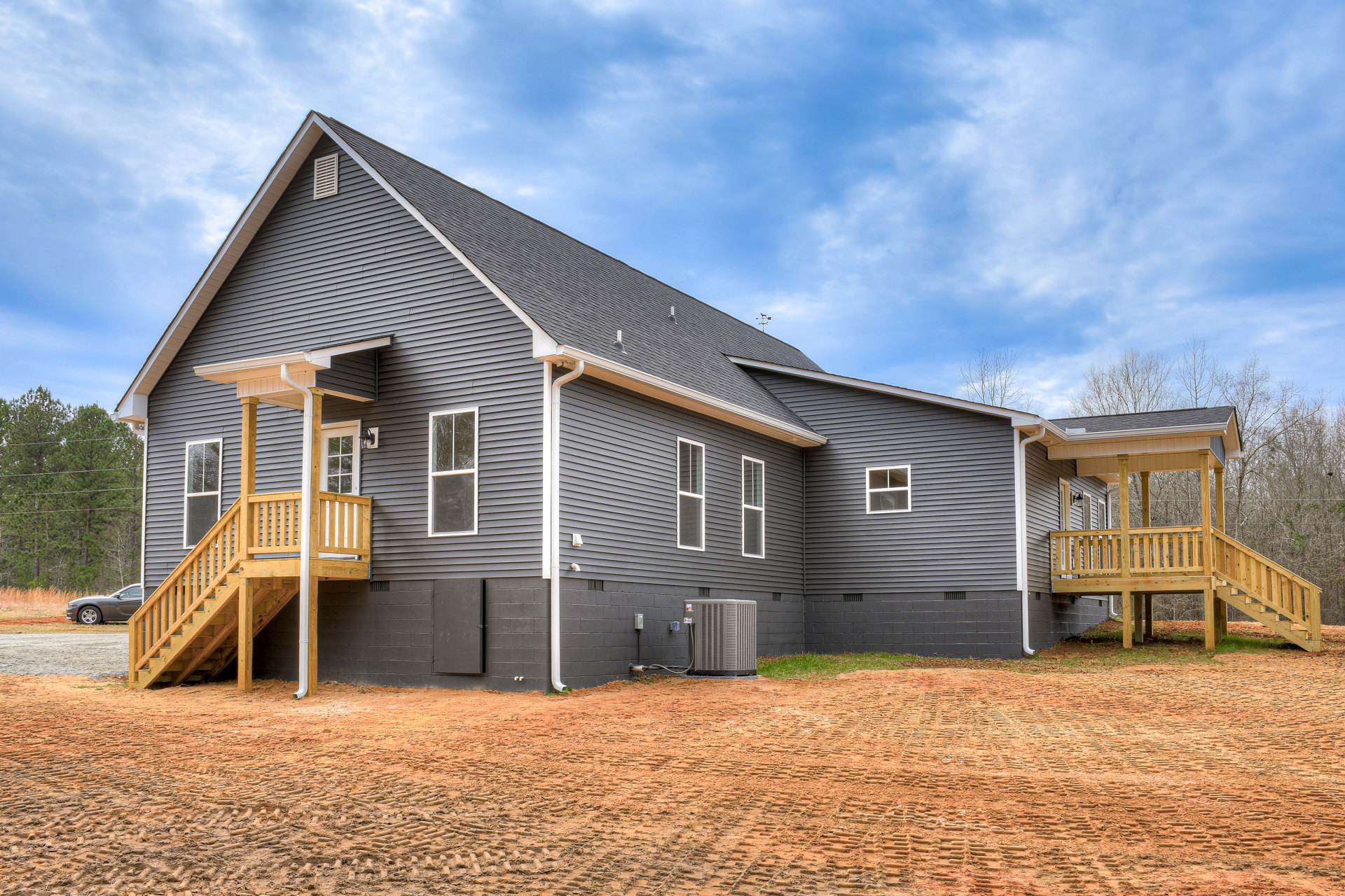 Two-story house with white-framed windows, wooden porch and stairs, gray siding, car parked beside gravel driveway, small machine on lawn, surrounded by trees under cloudy sky