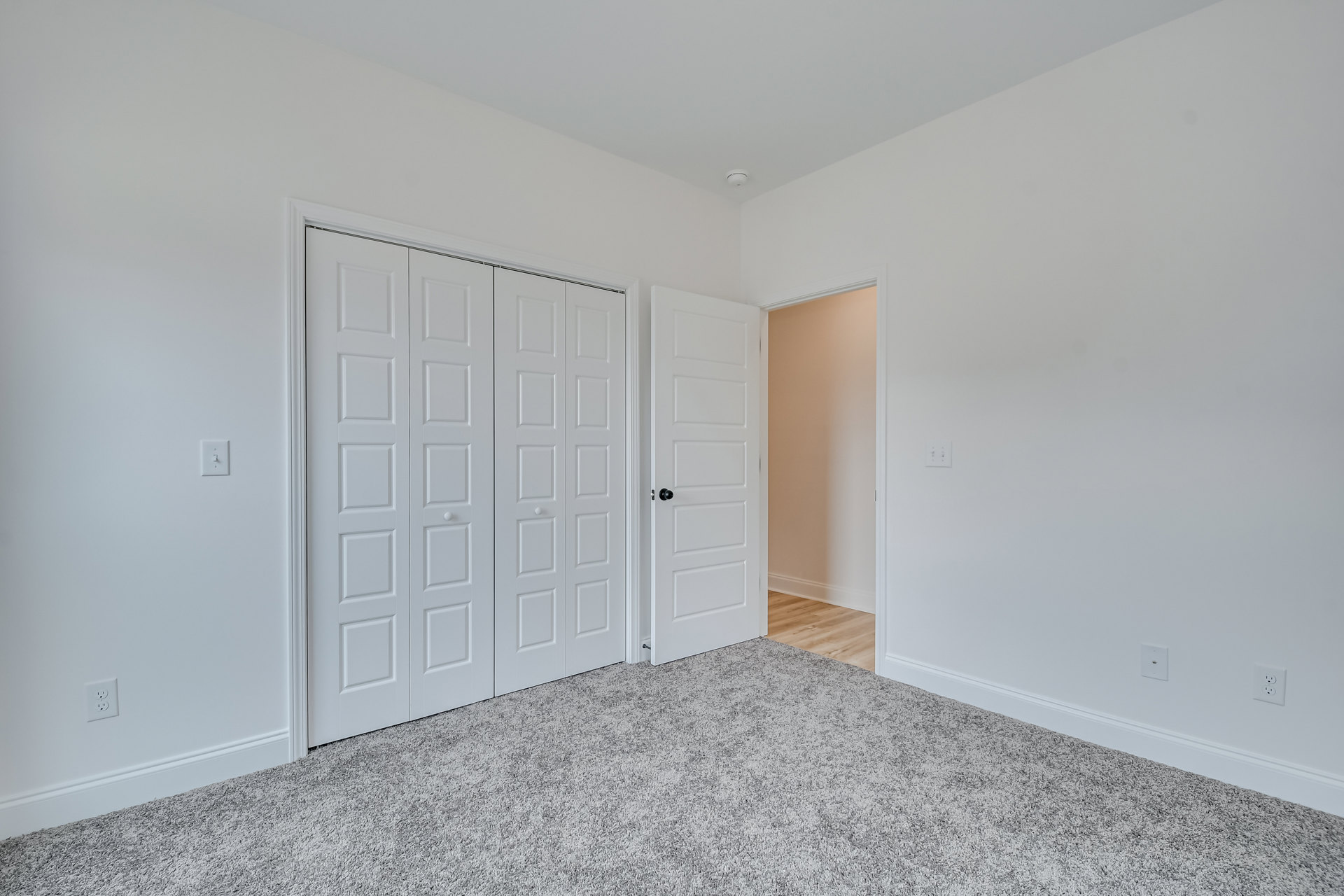 White walls and ceiling, white closet doors with black knobs, beige carpet flooring, simple trim and door frames